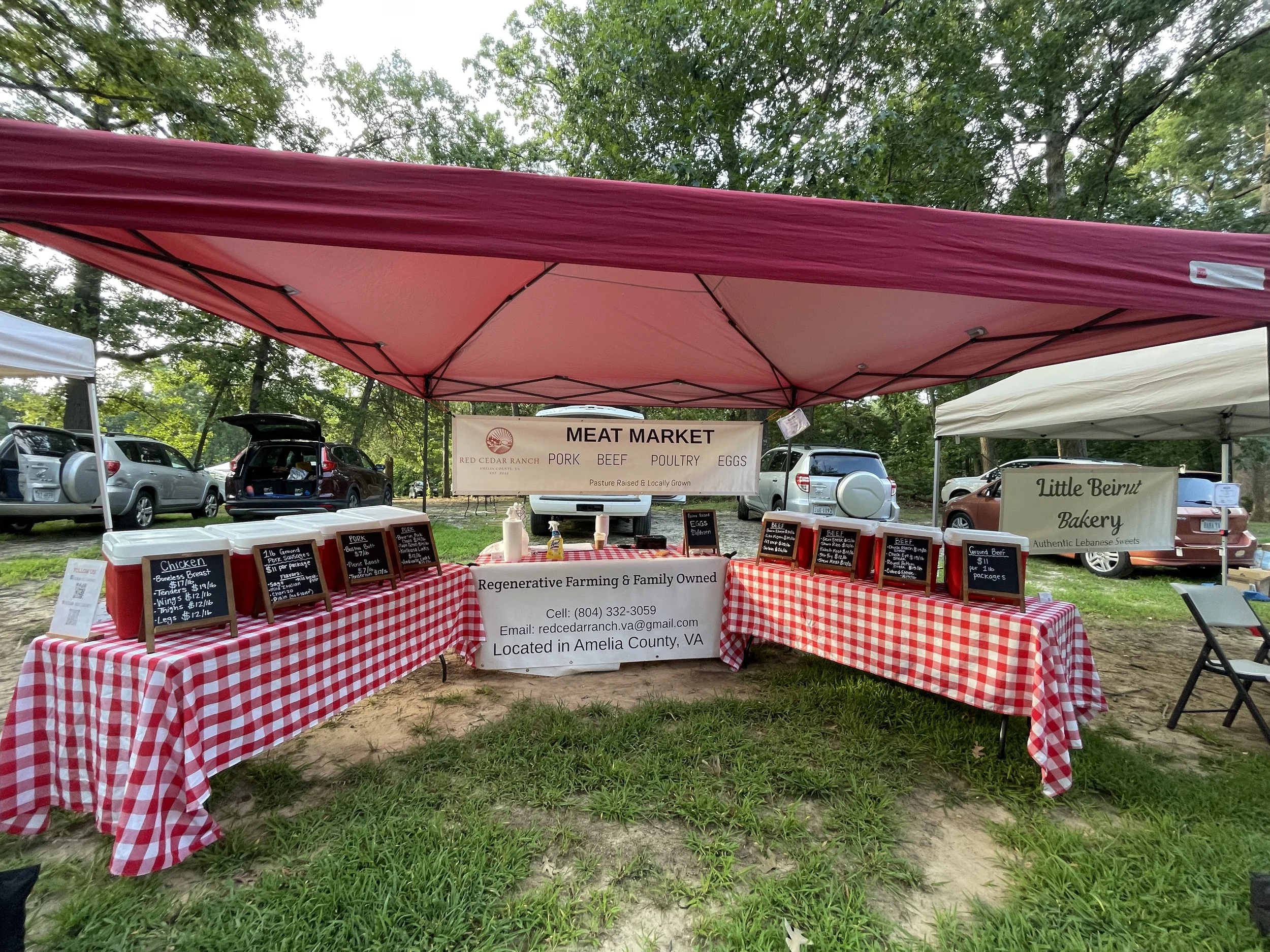Outdoor market stall with a red and white checkered tablecloth selling meat products. Sign behind the stall reads 'MEAT MARKET' with types of meat listed, such as pork, beef, poultry, and eggs. Multiple vehicles parked in the background among trees.