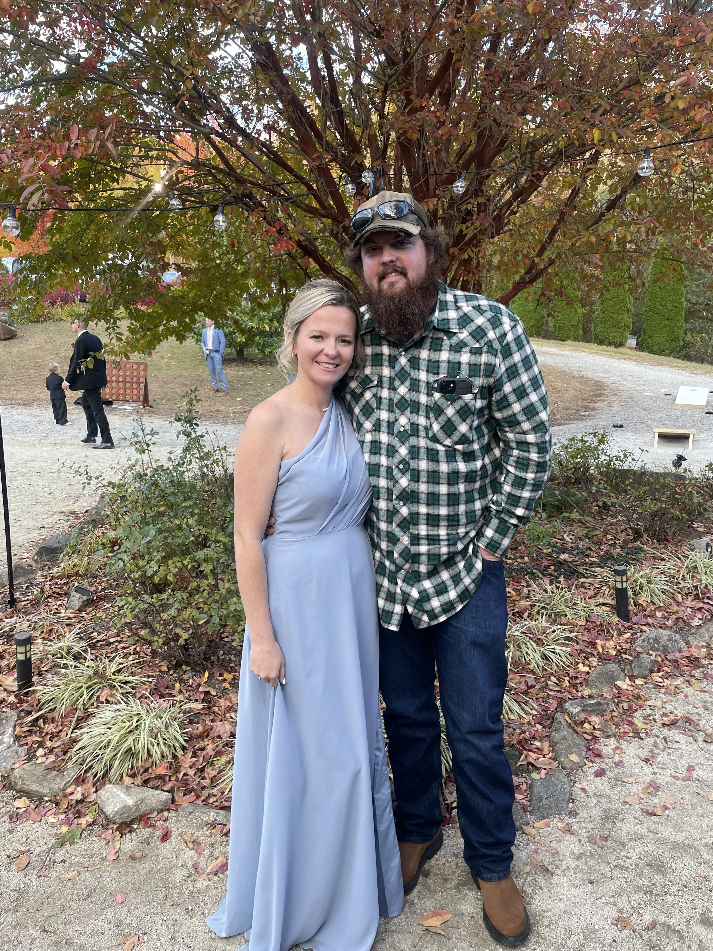 A woman in a light blue dress and a man in a plaid shirt stand together outdoors near a tree with red and green leaves. The scene appears to be at a festive gathering or outdoor event during autumn. The woman is smiling, and the man has a beard, wears sunglasses on his cap, and has a camera in his shirt pocket. Other people are seen in the background.