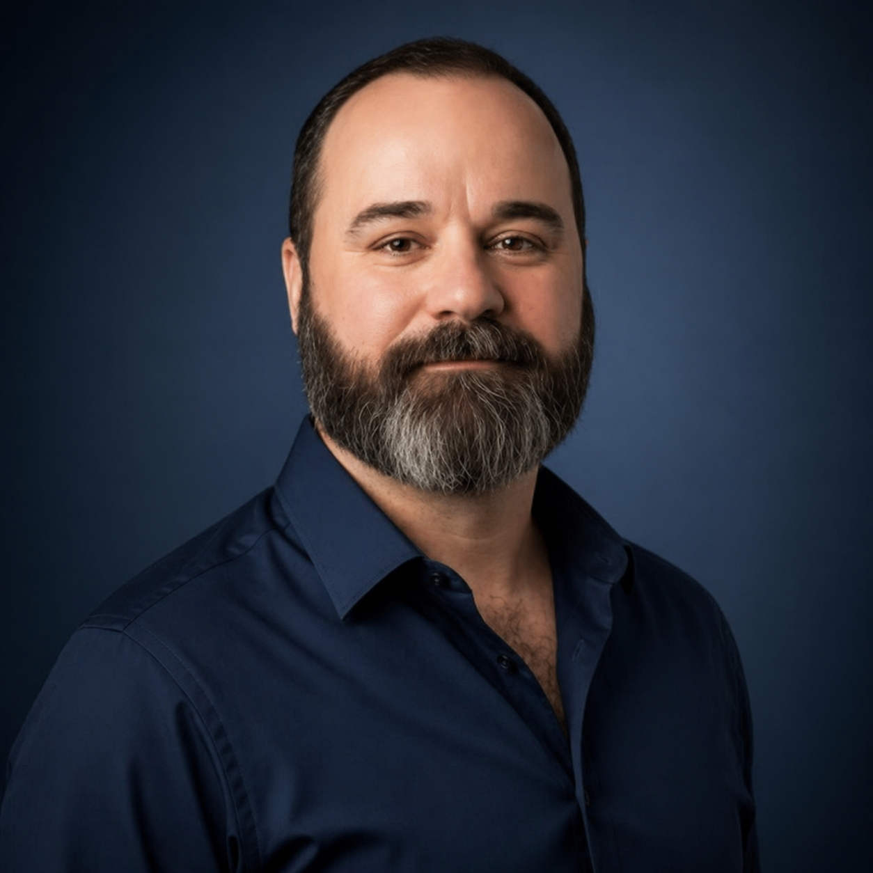 A professional headshot of a man with a beard, wearing a dark blue collared shirt, against a dark blue background.