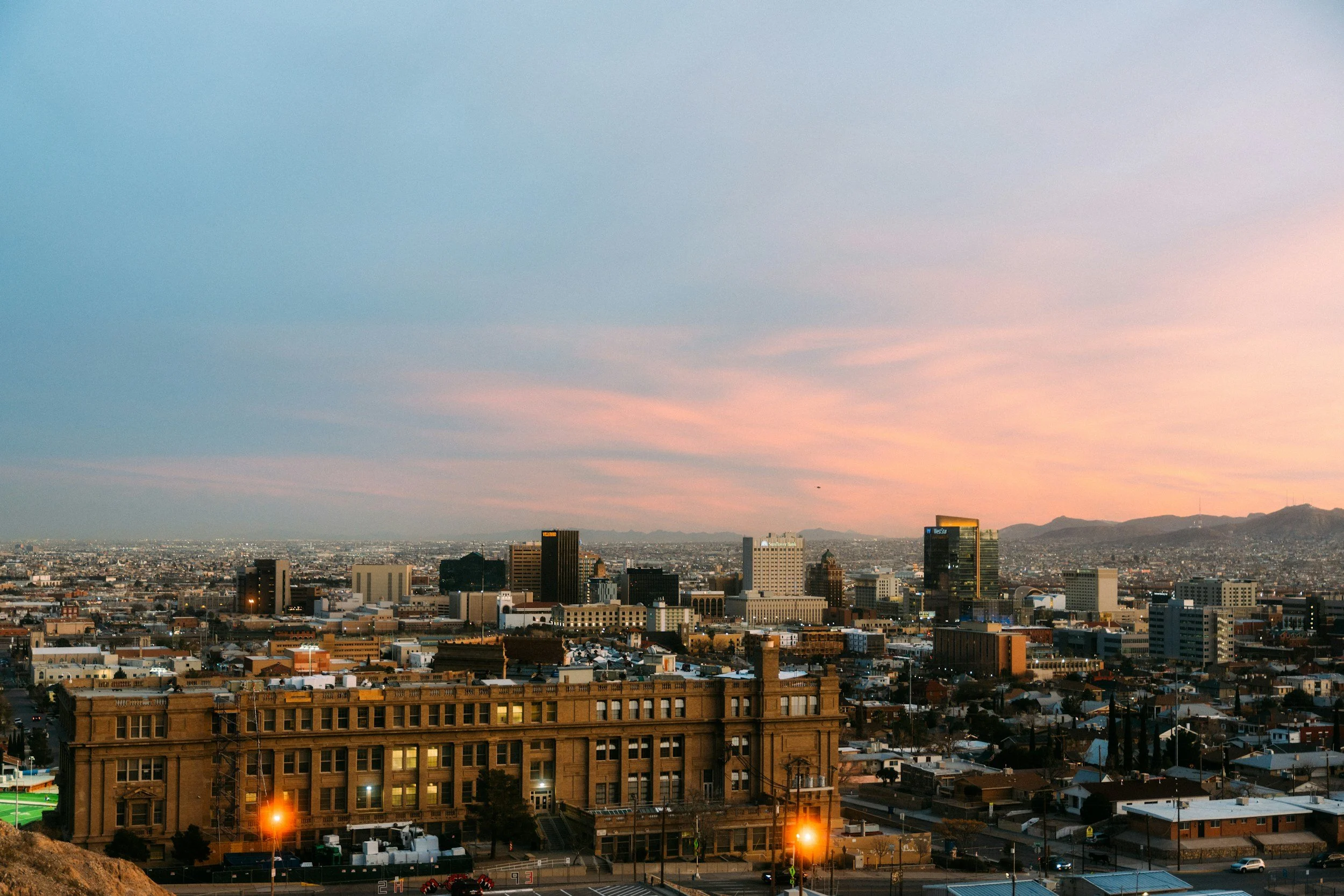 A cityscape view during sunset with numerous buildings, including skyscrapers, under a colorful sky with shades of pink and blue.