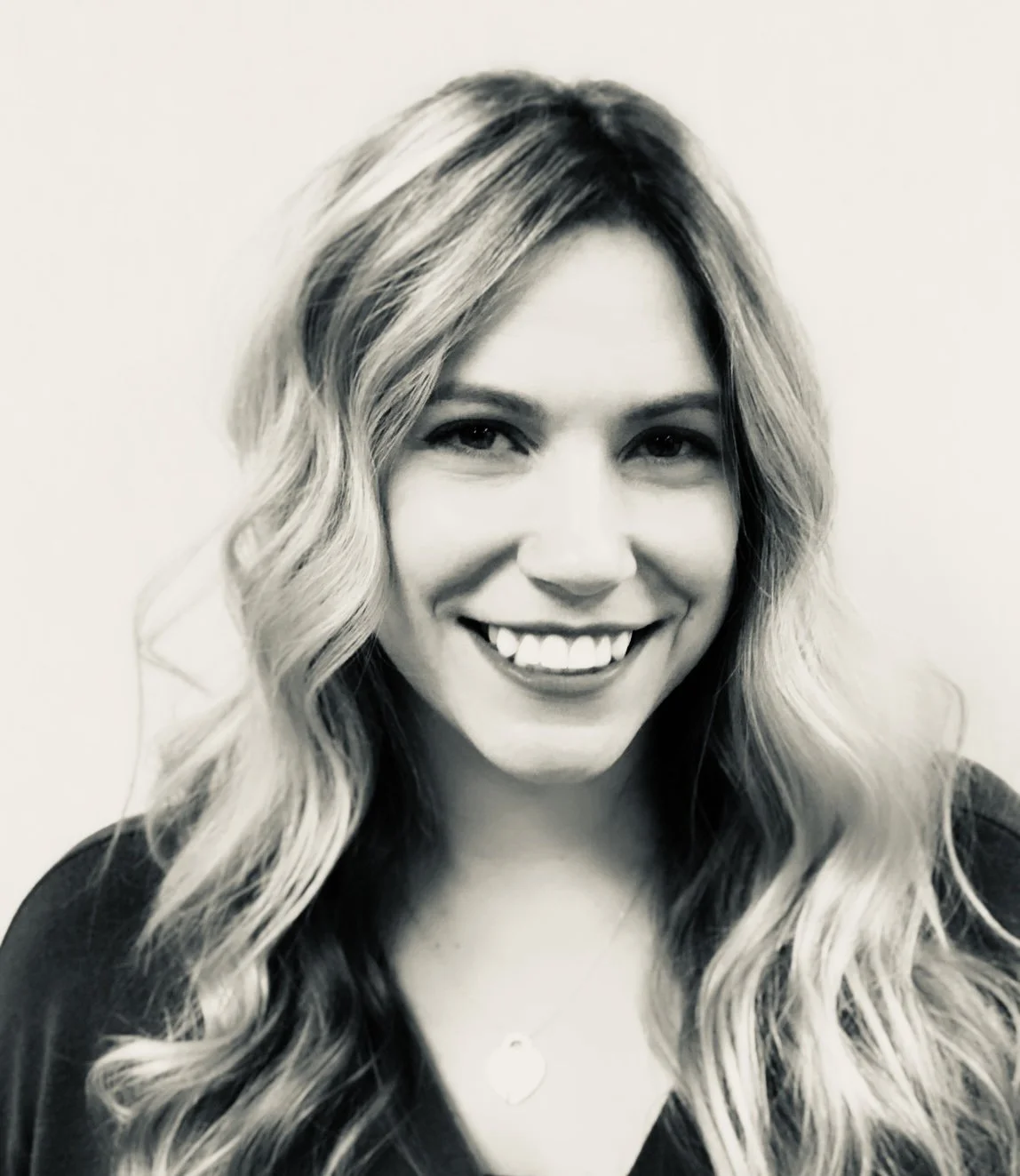 Black and white photo of Abbey Lynne Rays, smiling, with long, wavy hair, wearing a necklace and a dark top.