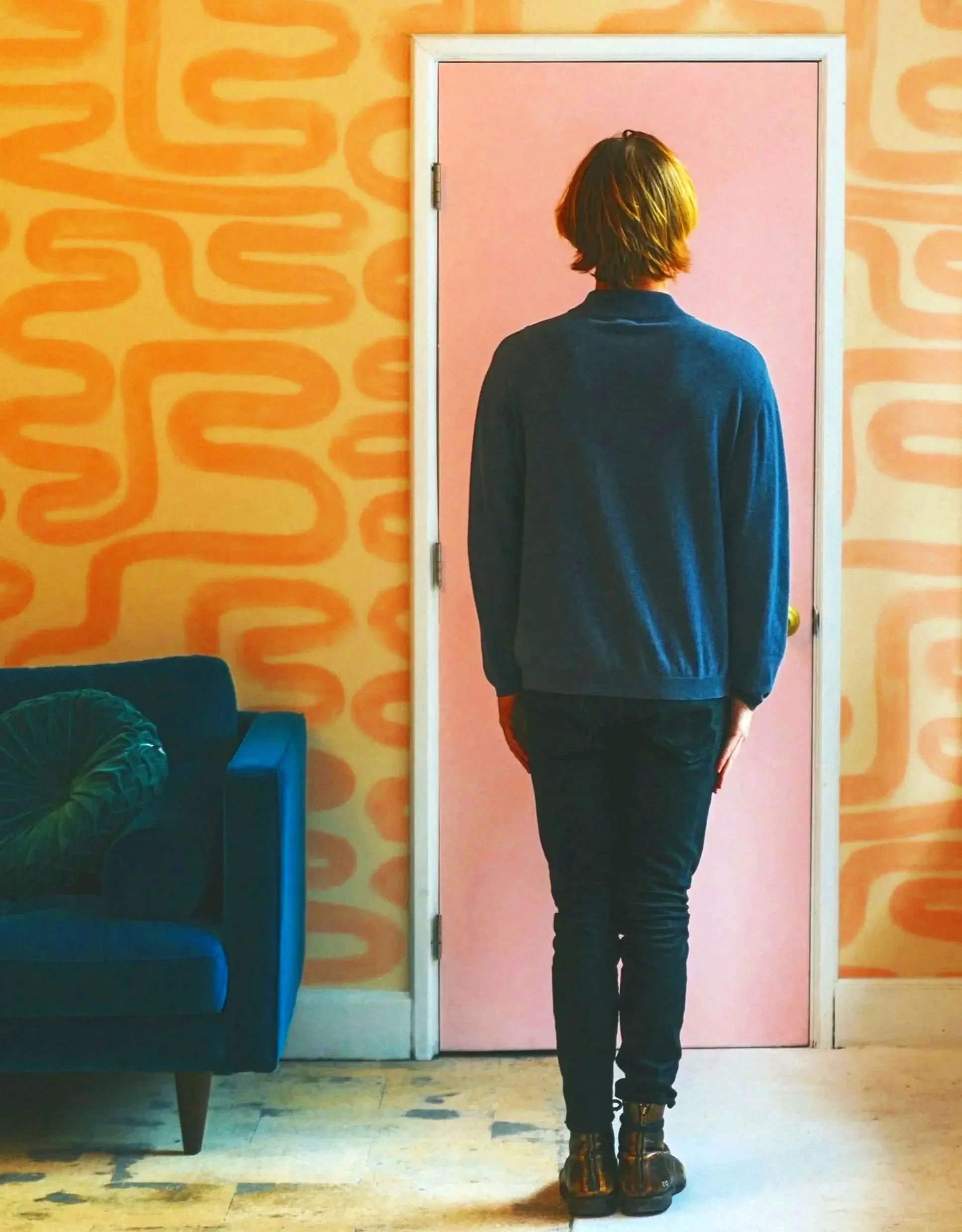 A person stands facing a pink door in a room with bright orange patterned walls.