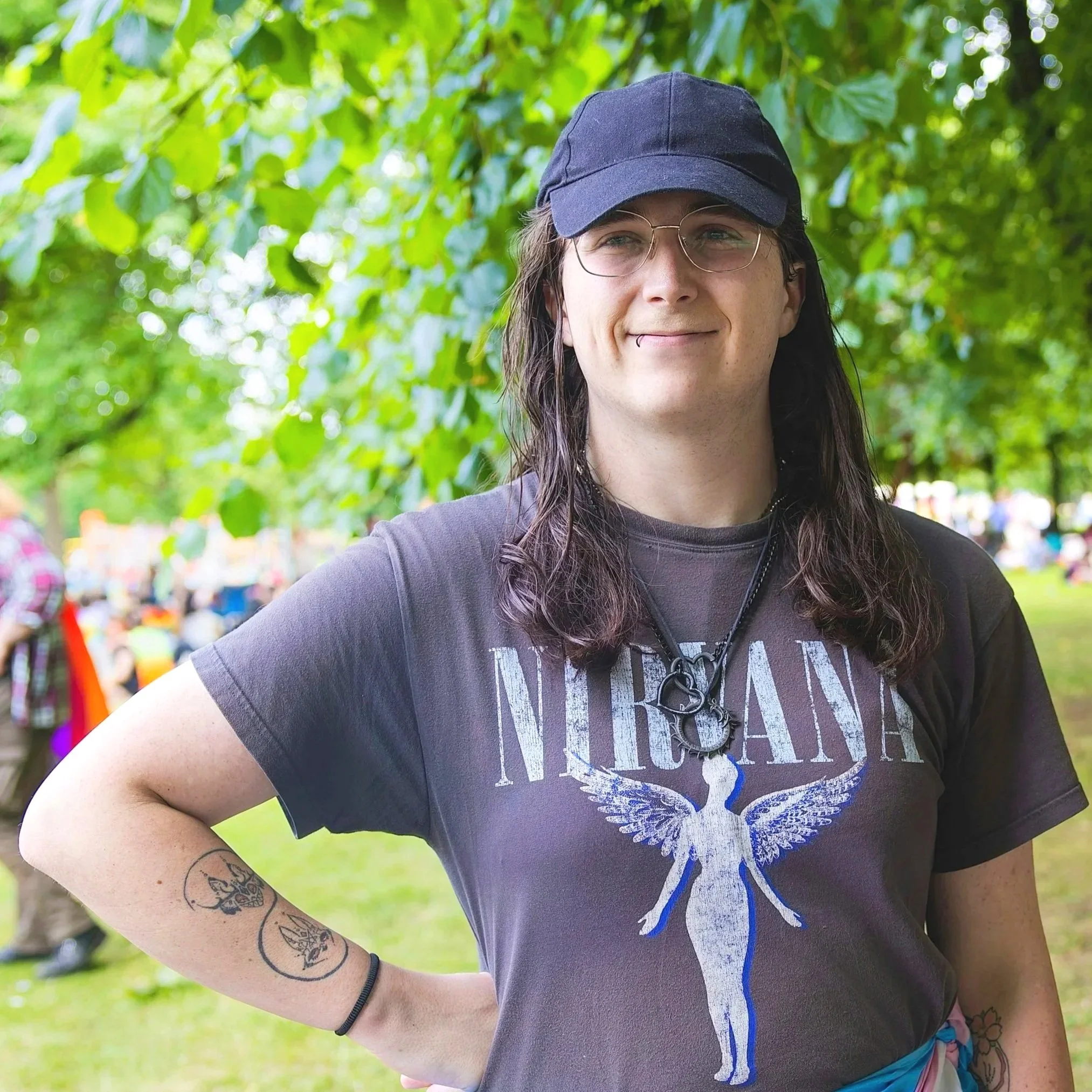 A trans woman with long dark hair, glasses and a baseball cap stands outdoors beneath green trees, one hand on her hip and a warm, self-assured smile conveying confidence, ease and quiet happiness in who she is.