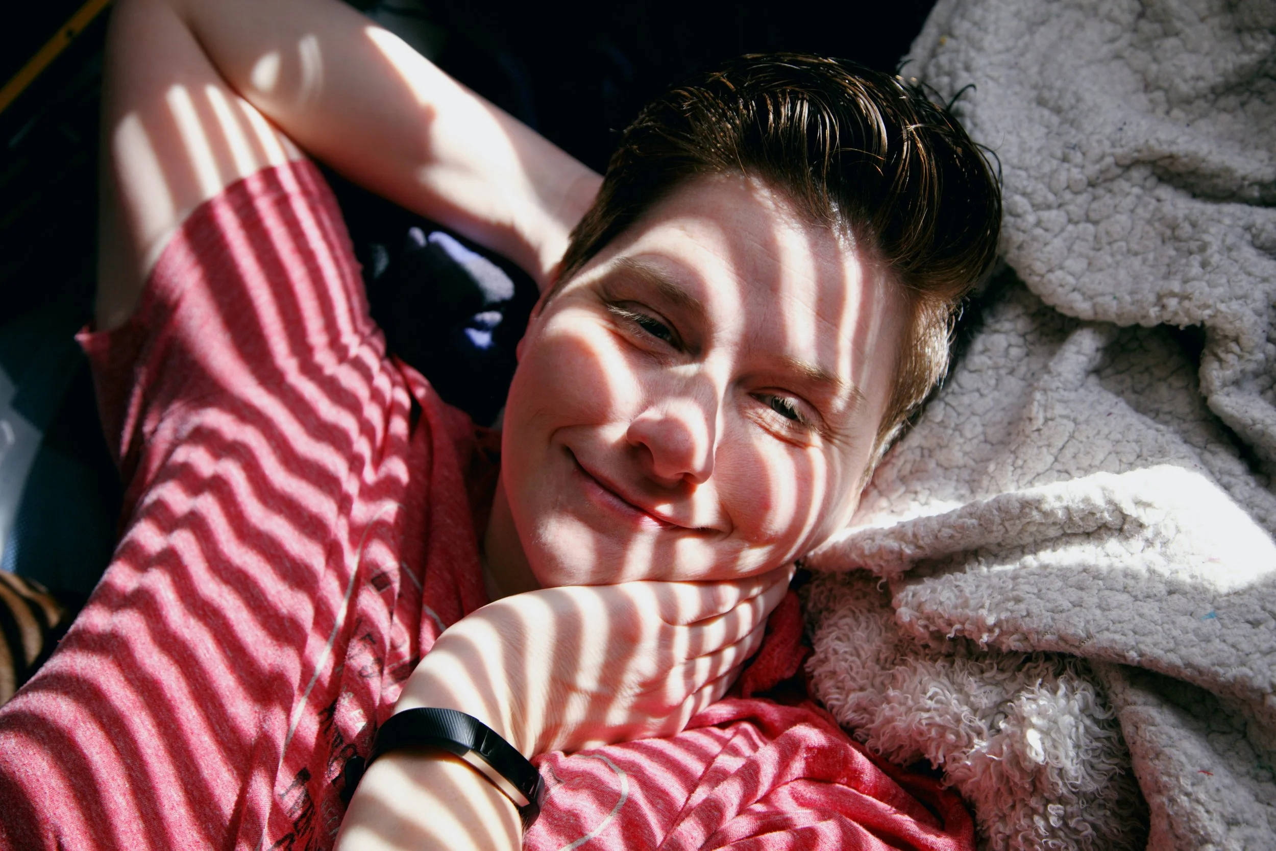 A person in a red shirt reclines against soft blankets as striped sunlight crosses their face and arms; their relaxed posture and gentle smile suggest ease, playfulness and greater access to pleasure.
