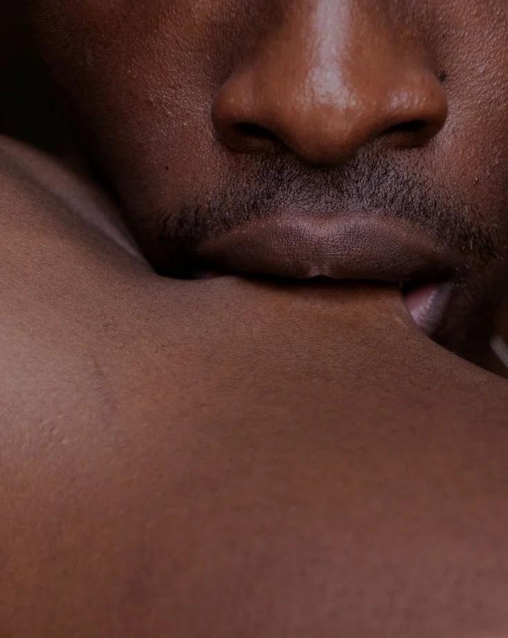Close-up of a Black person gently and tentatively tugging at bare skin with their teeth, their expression tender and intimate.