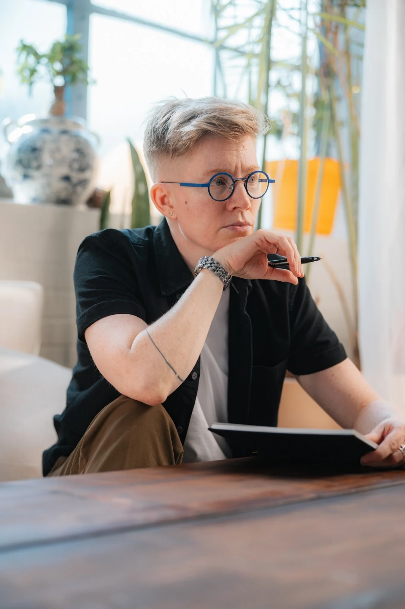 Ezra Cake holds a pen above an open notebook while listening to someone, their thoughtful expression and attentive posture conveying presence, curiosity and care.