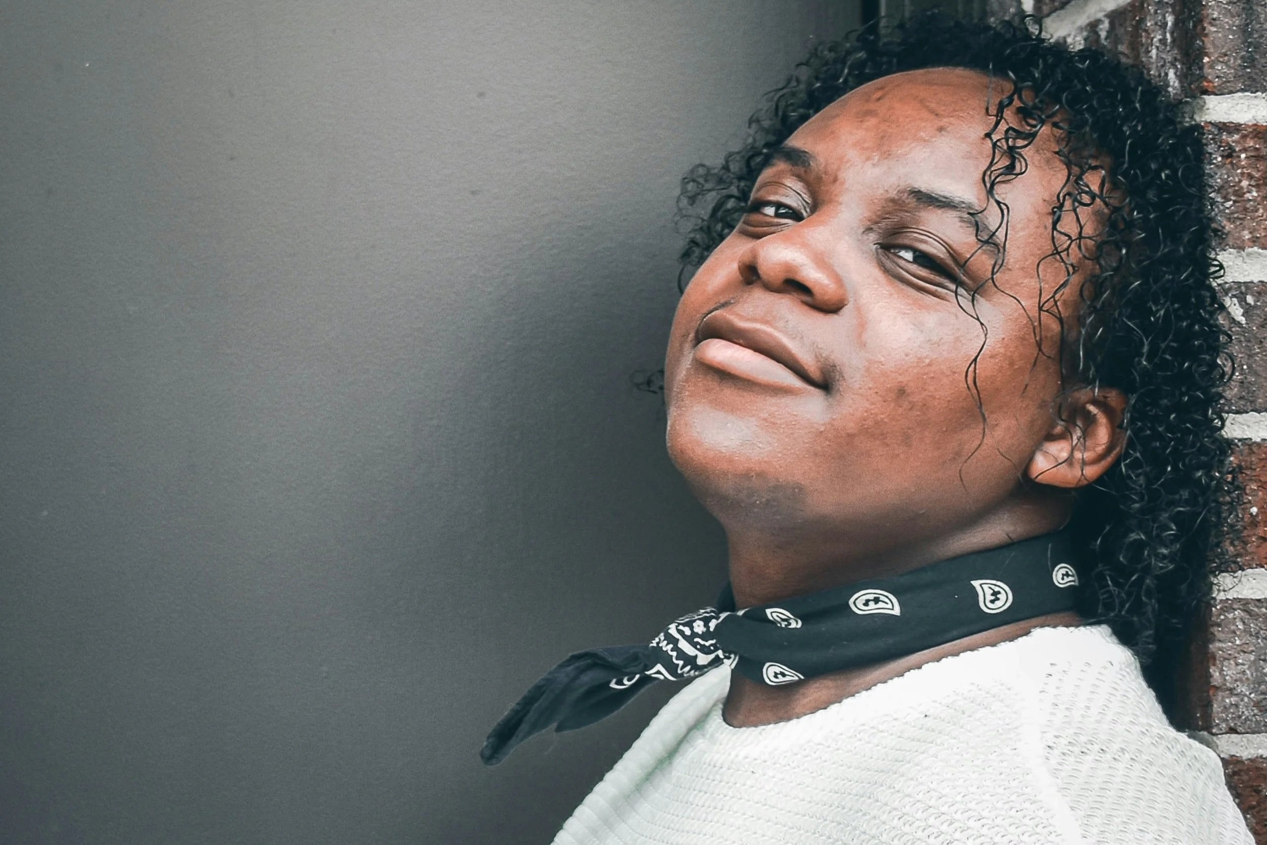 A Black gender diverse person with curly hair and a dark bandana leans against a brick wall, gazing upward with a soft, self-assured smile that conveys hope, ease and self confidence.