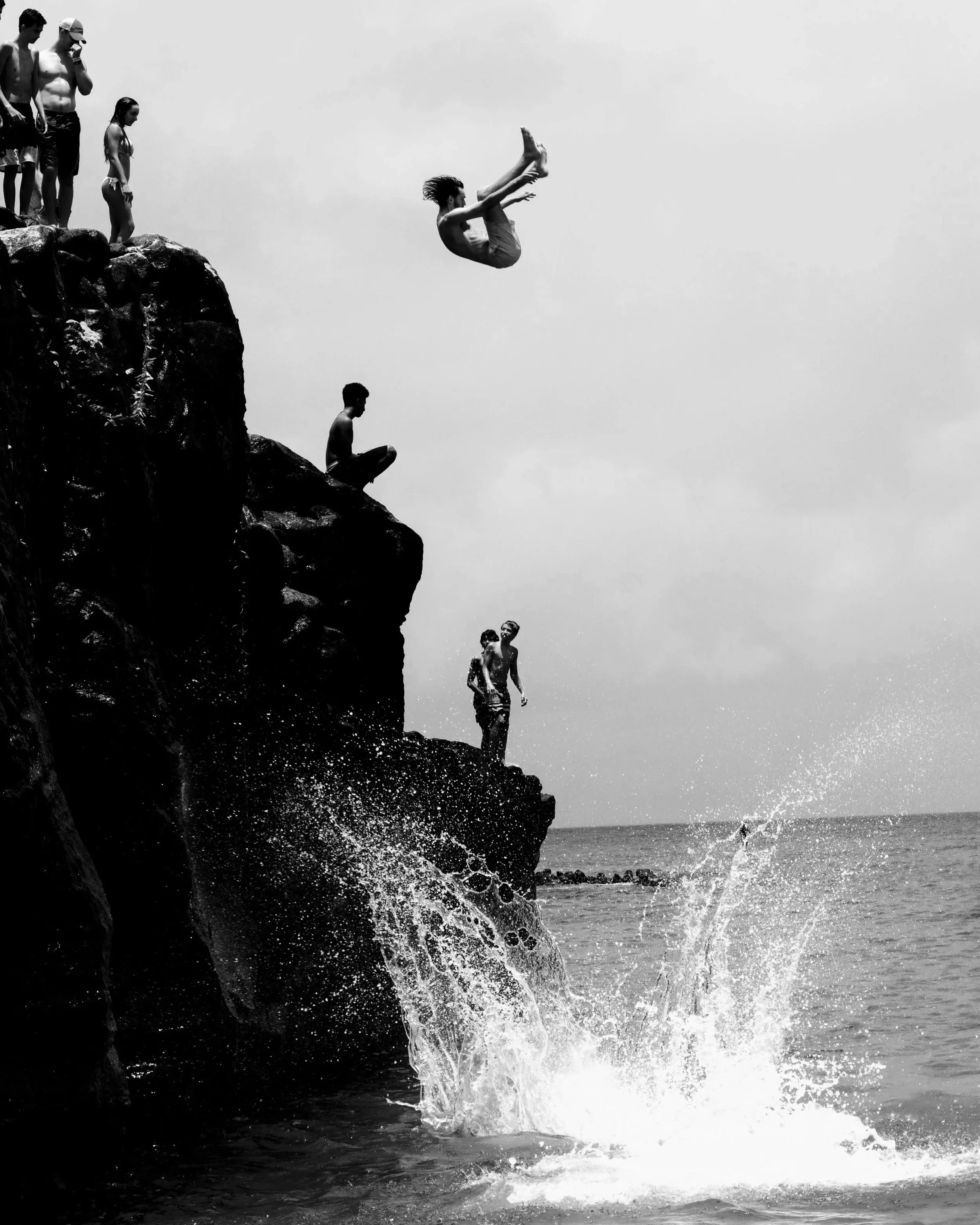 Black-and-white photograph of a person flipping from a rocky cliff as others watch, with a large splash below.