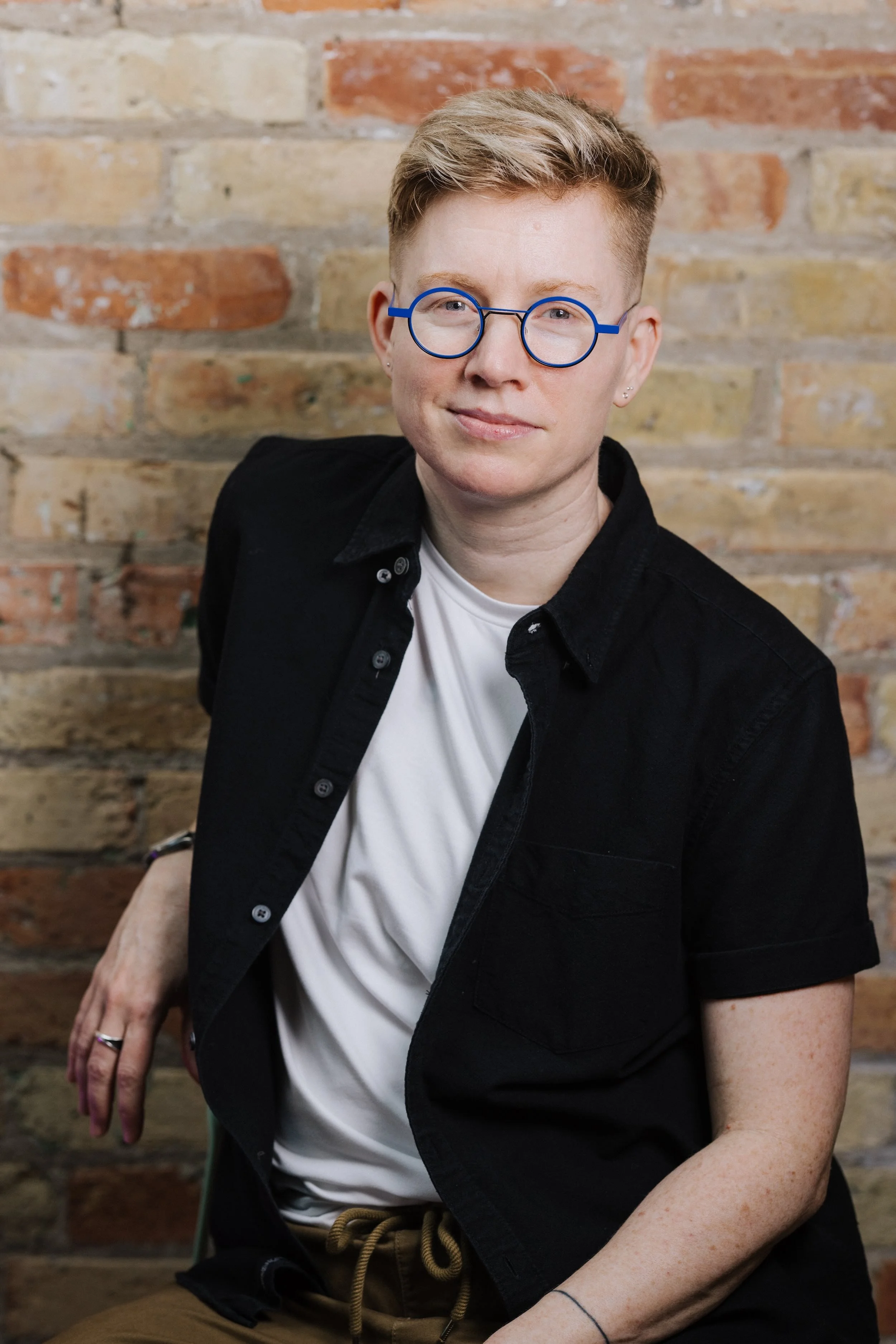 Ezra sits against a warm brick wall, wearing bright blue glasses and meeting the camera with a calm, direct expression.