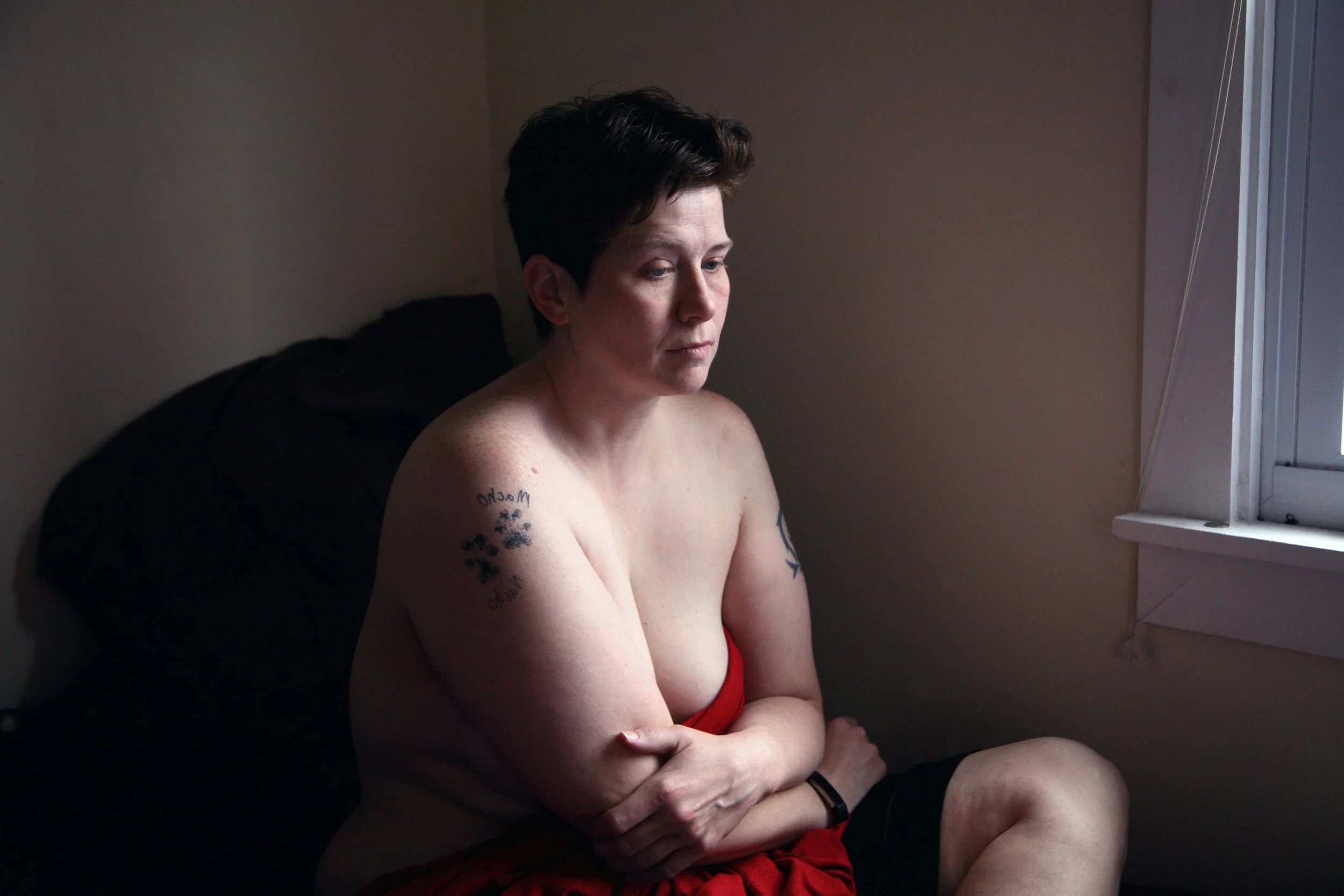 A bare-chested person with short dark hair sits wrapped in a red blanket, arms folded around their body and gaze lowered, conveying vulnerability, self-protection and the emotional weight stress around sexuality can carry.