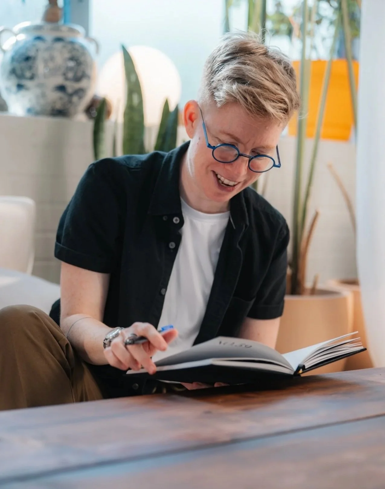 A young man with blond hair and glasses, sitting indoors, smiling and reading a book.