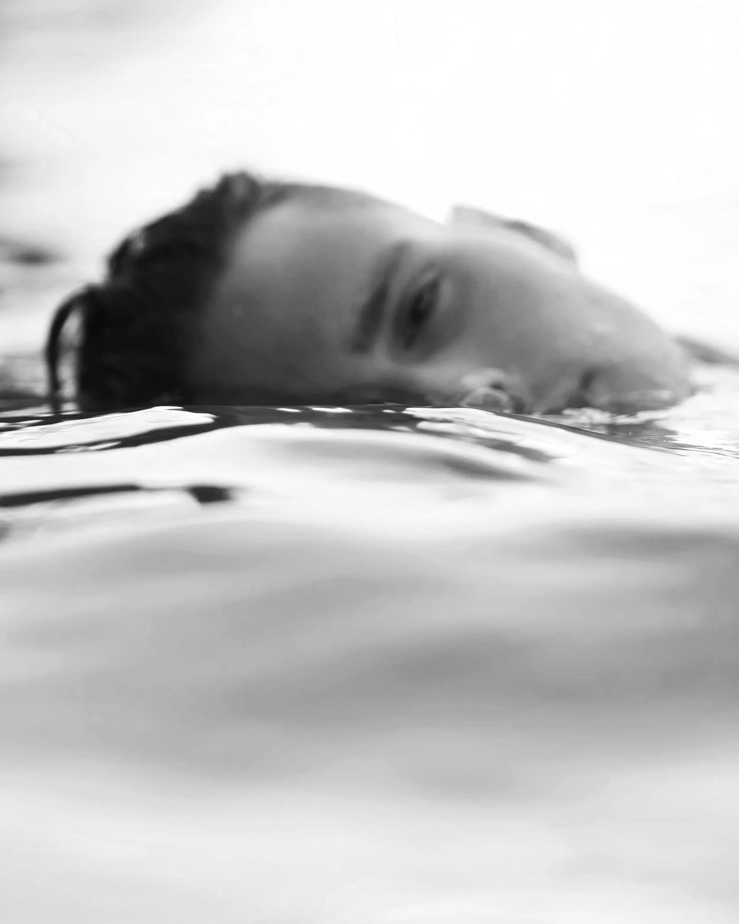 A gender-ambiguous person rests their face at the surface of the water, their reflection rippling around them.