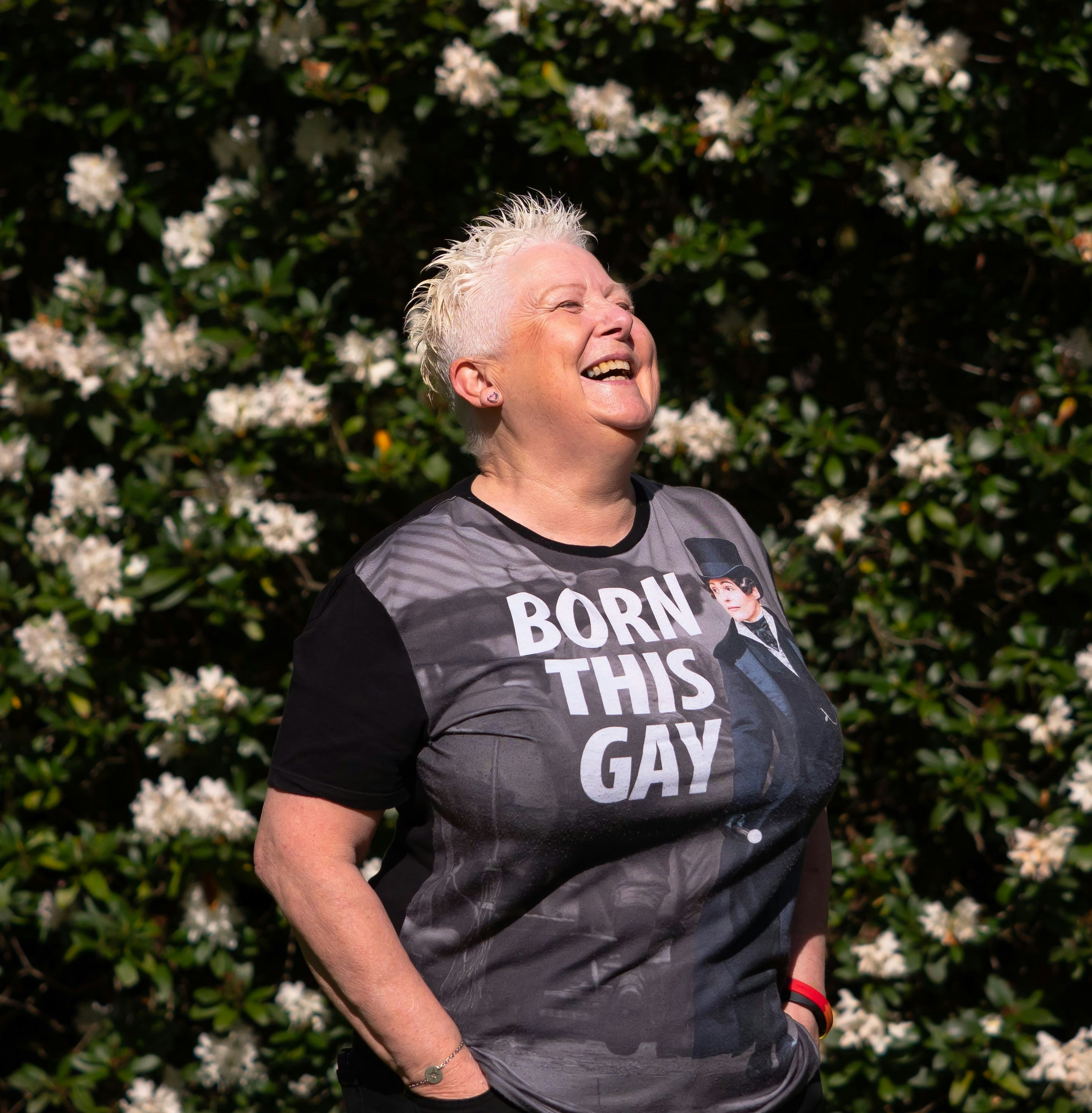 An older person laughs outdoors among flowering bushes, wearing a shirt that reads “Born This Gay.”