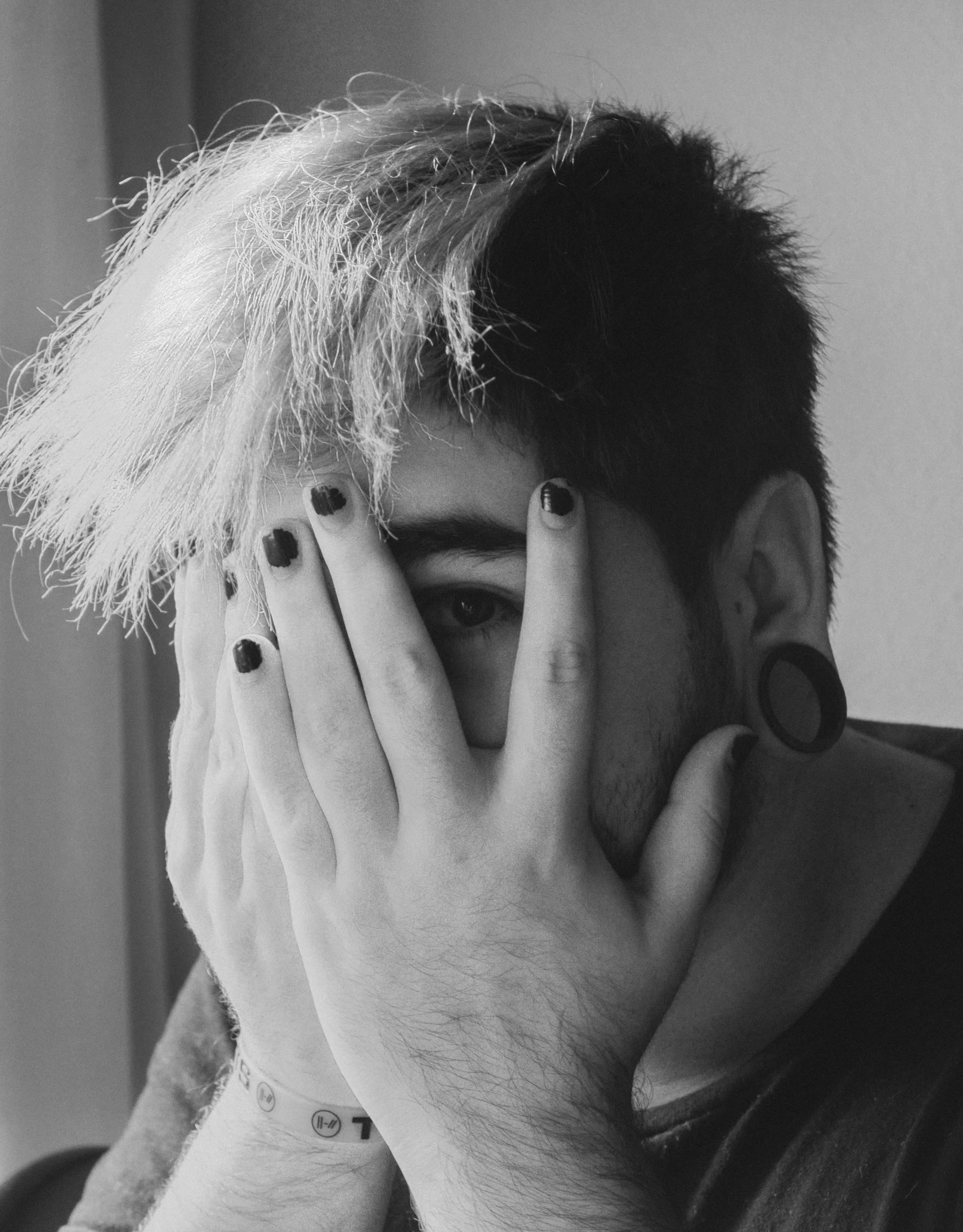 A person with short hair, painted fingernails and stretched earlobes looks through their fingers while covering most of their face.