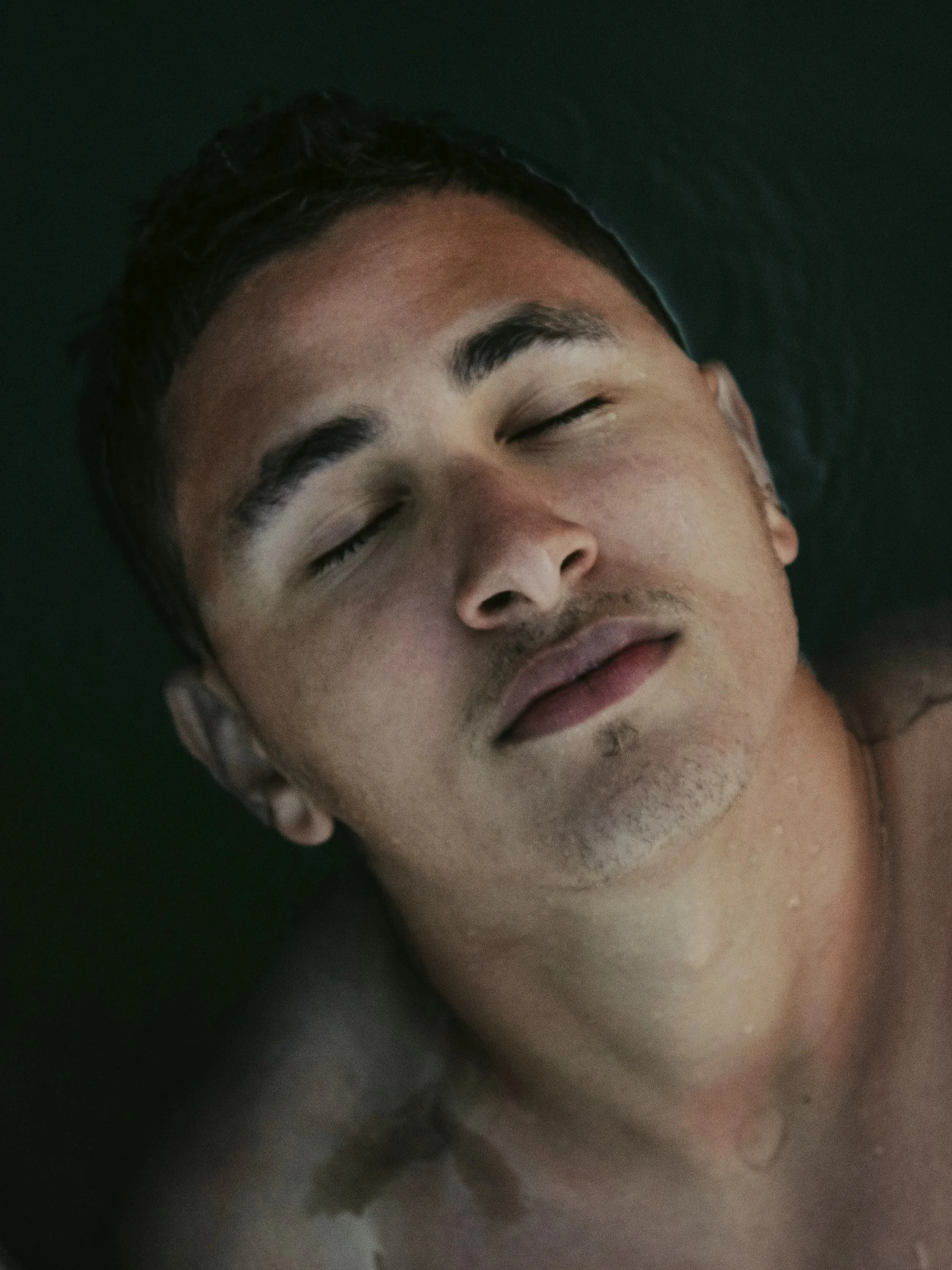 A person floats partially submerged in dark water with their eyes closed, appearing peaceful, absorbed in pleasure and at ease.