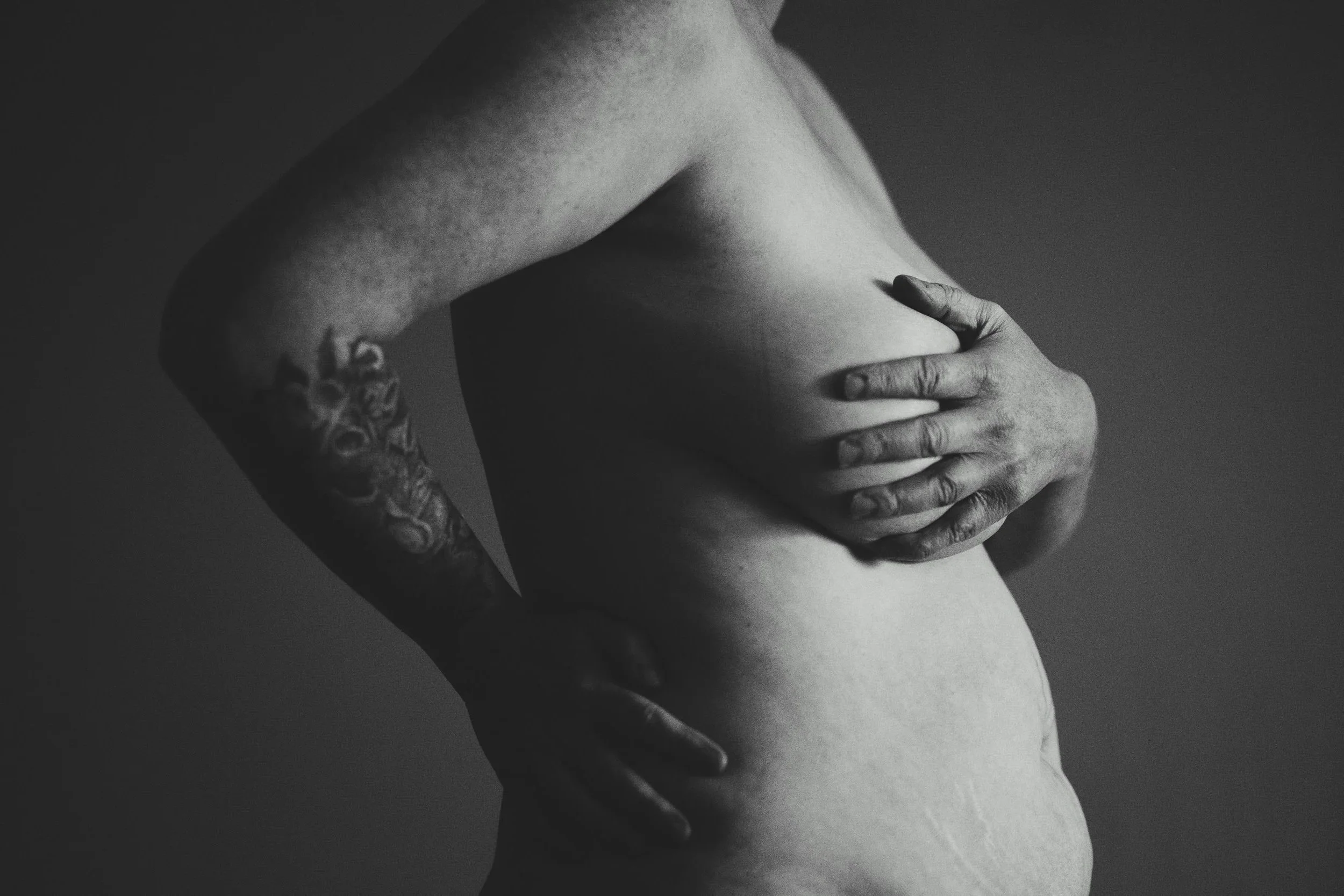 Black-and-white close-up of a middle-aged person holding their bare torso, with stretch marks and a floral tattoo visible; the touch feels protective and uncertain.