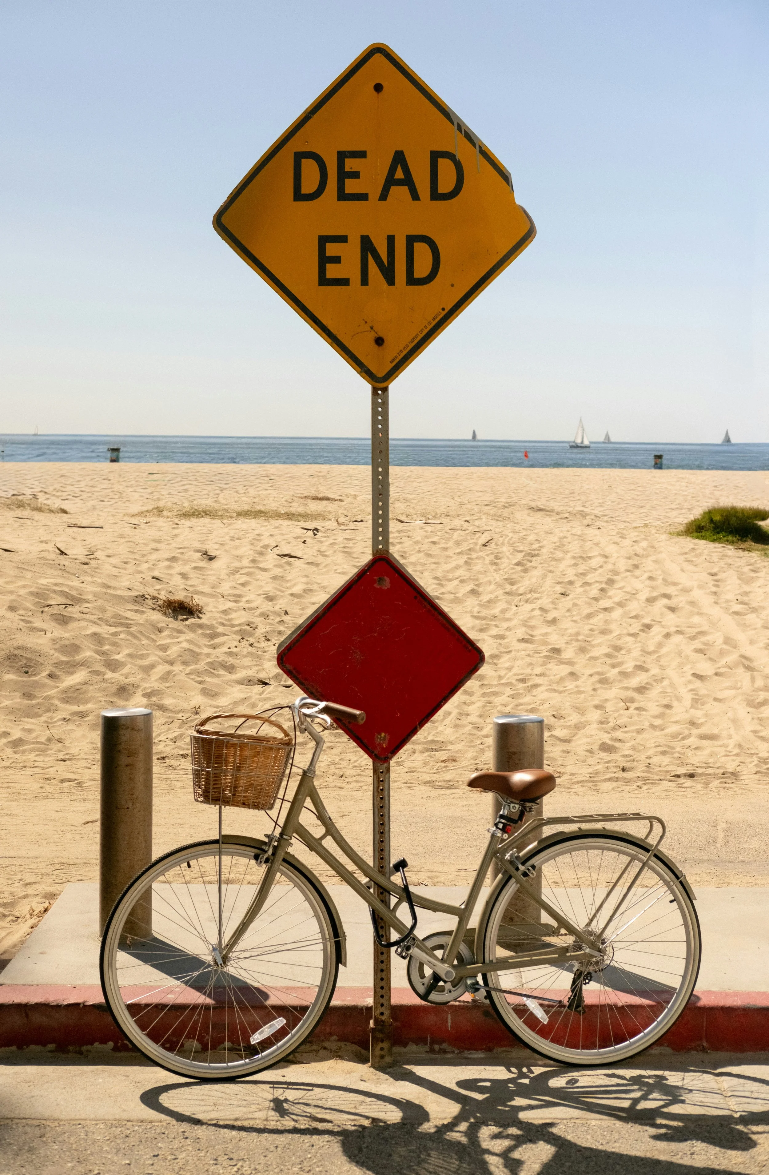 A yellow “DEAD END” road sign stands on a sandy beach, with the water and sailboats behind it and part of a bicycle below.