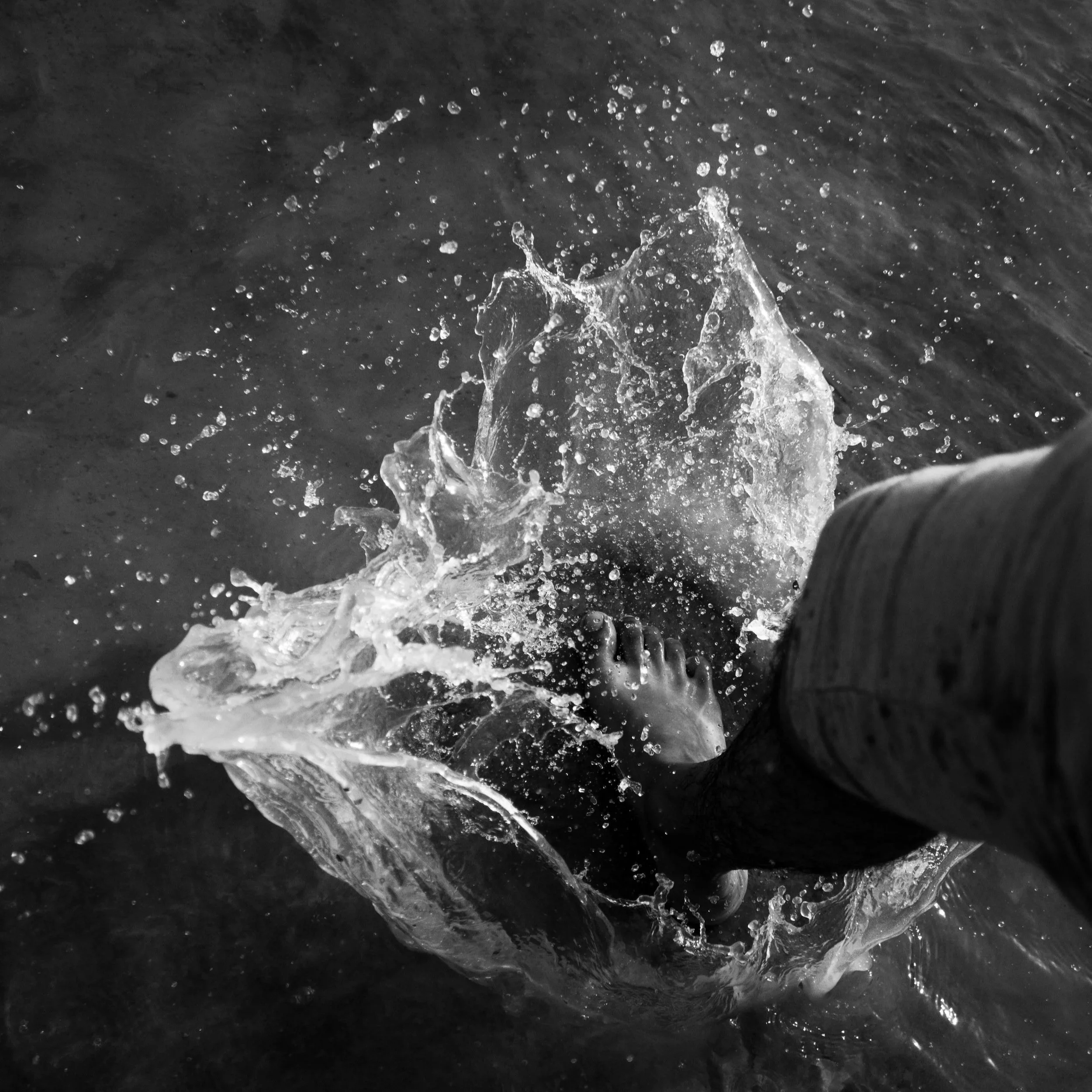 Black-and-white overhead view of a person plunging their foot into dark water, surrounded by a dramatic burst of spray.