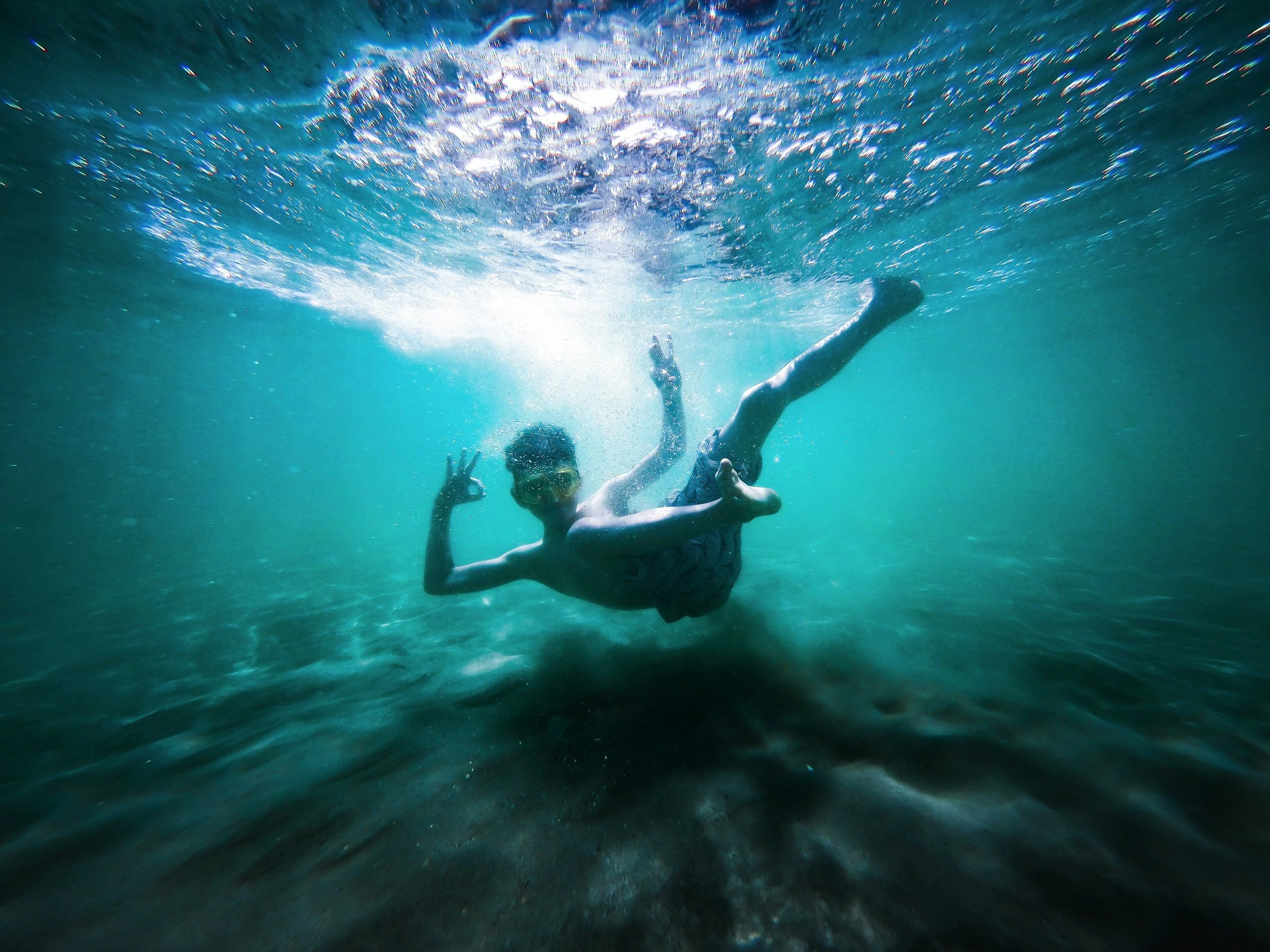 A swimmer floats freely beneath the sunlit surface of clear blue water, making an “okay” sign with one hand, their body loose and playful above the rocky seabed.
