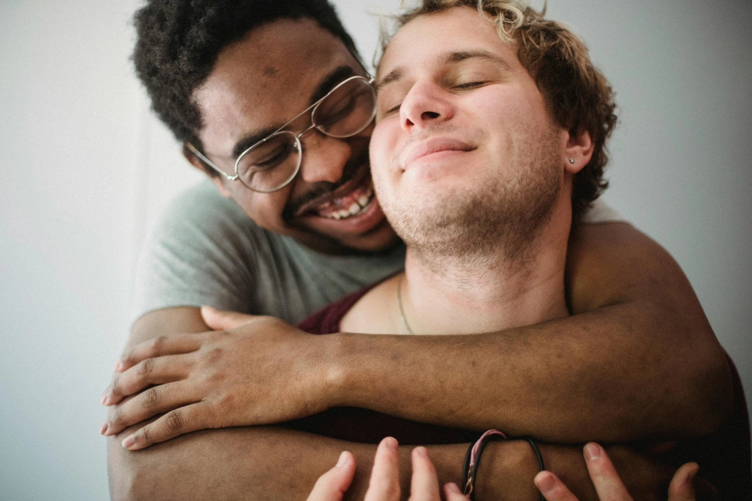 A Black man wearing glasses smiles joyfully as he wraps his arms around a white man with an earring, who closes his eyes with a relaxed smile; their close embrace conveys intimacy, trust and ease in connection.