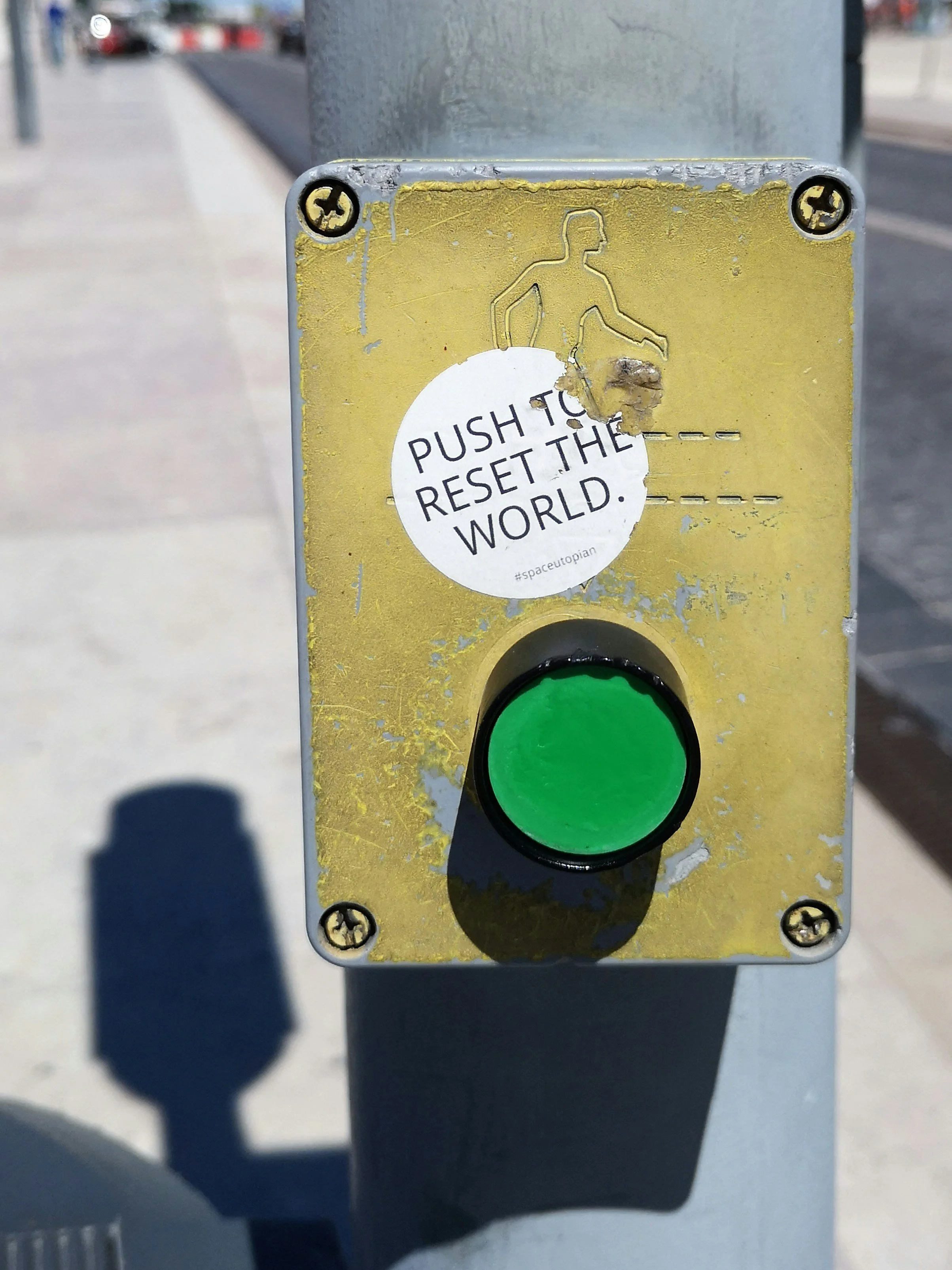 A green button on a weathered yellow pedestrian crossing box, with a sticker above it reading “Push to reset the world.”