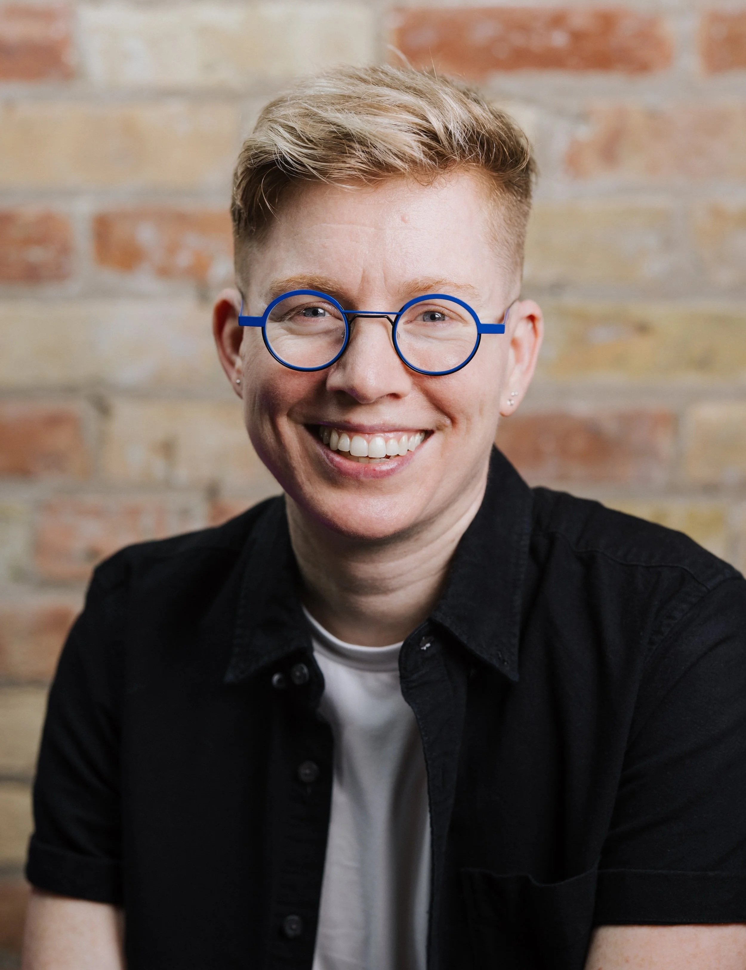 Close-up portrait of a smiling person with short blond hair, wearing round blue glasses, a white t-shirt, and a black shirt, standing against a brick wall background.