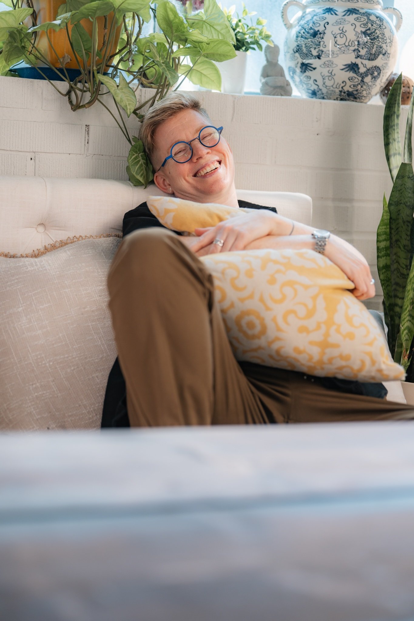 Ezra Cake with short hair and glasses, smiling, joyful, relaxed, leaning back hugging a patterned yellow and white pillow, surrounded by green plants and decorative objects in a cozy space.
