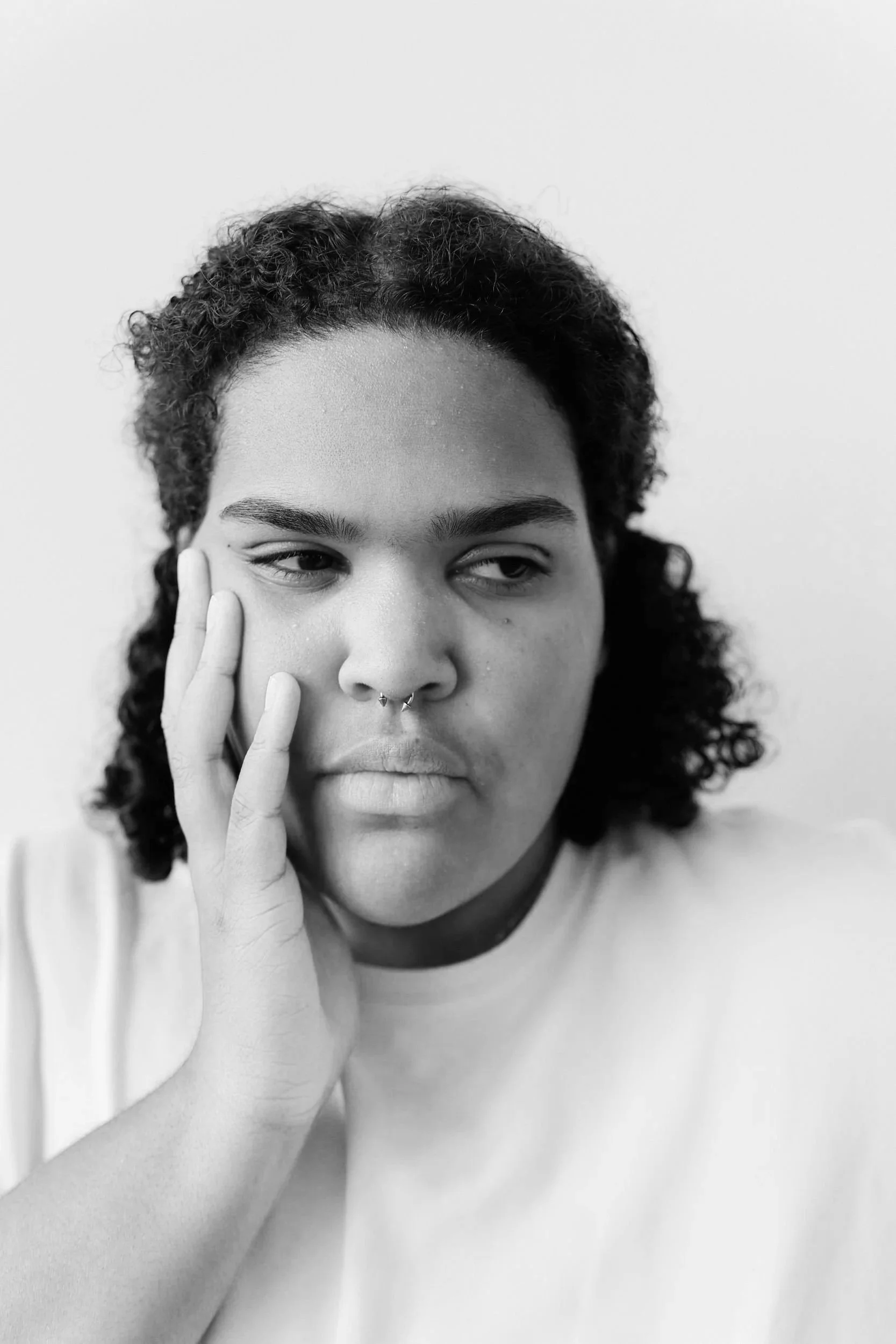 A Black person with curly hair and a septum ring rests their cheek in one hand and looks to the side.