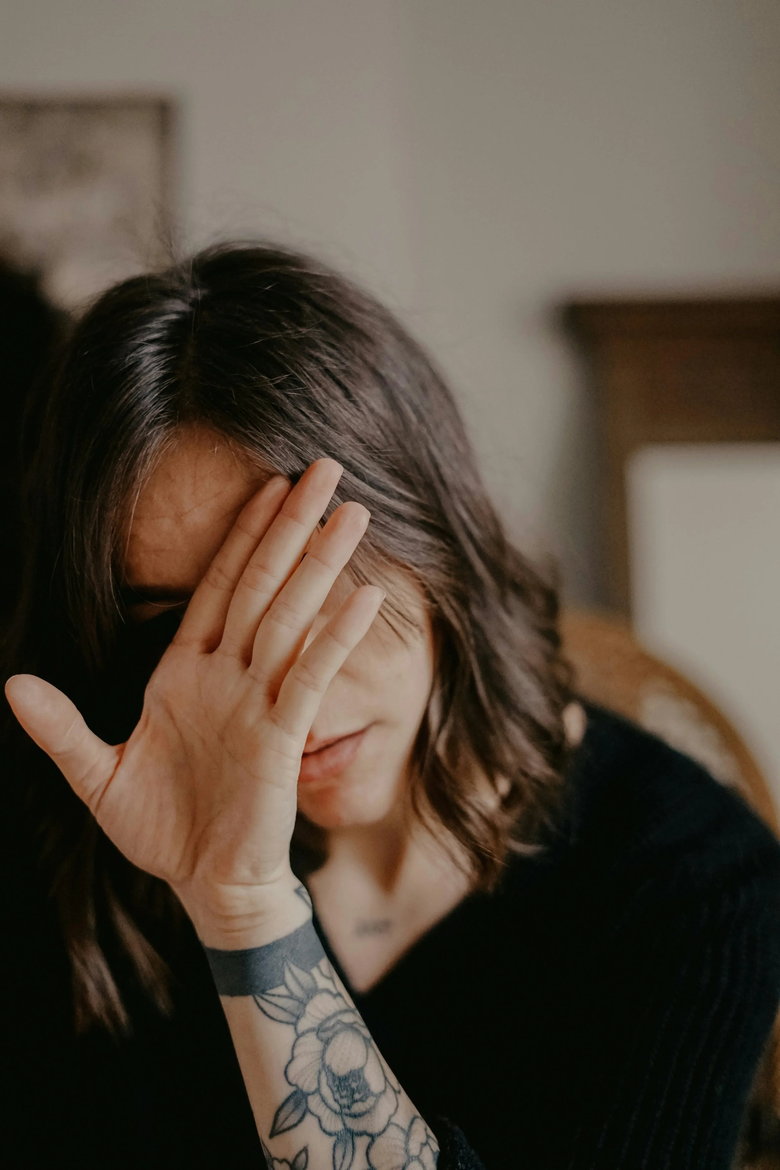 A trans woman with long dark hair lowers her head and raises one hand to shield her face, her partially hidden expression and closed posture conveying discomfort, hiding and self-protection.