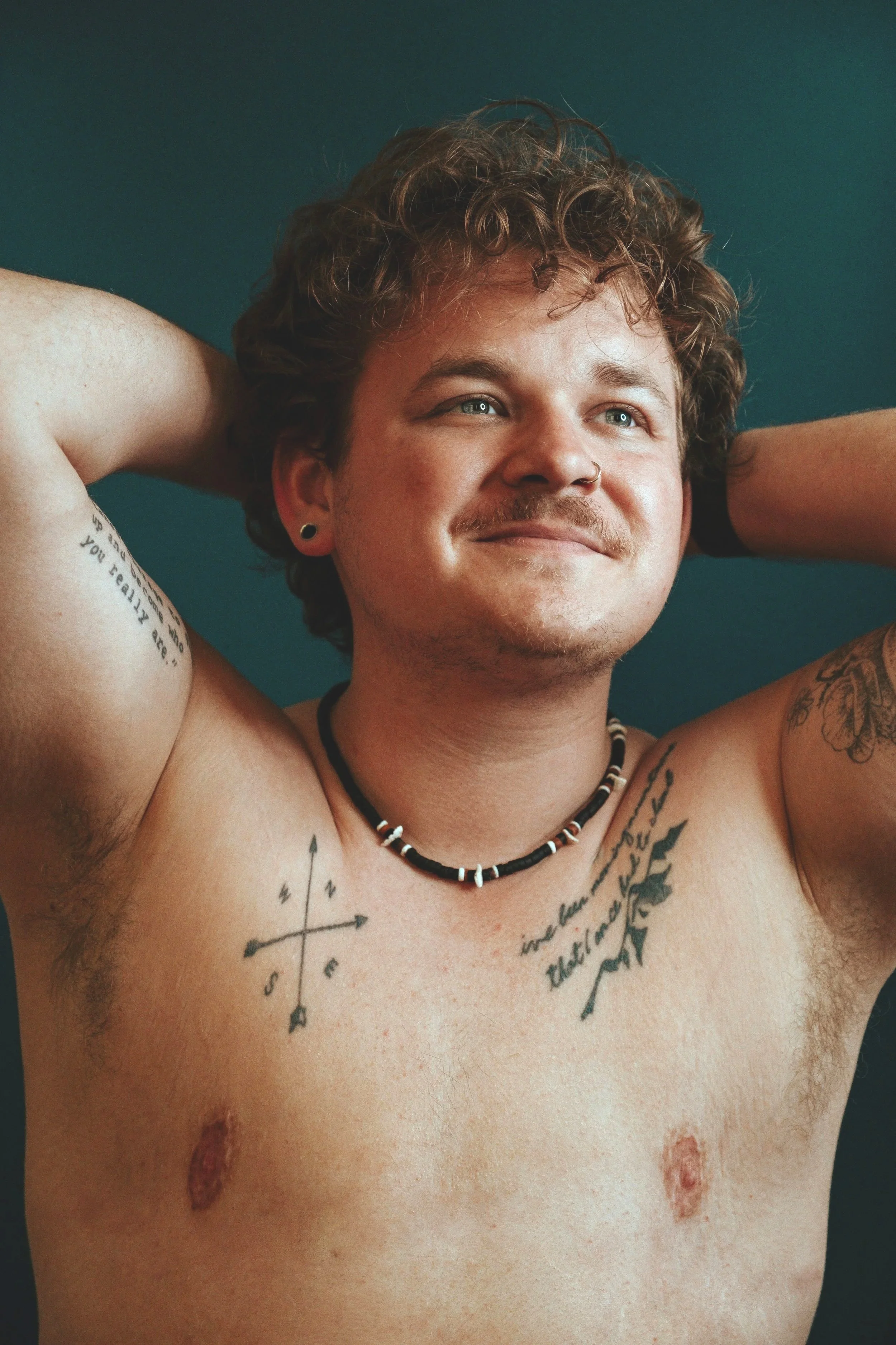 A bare-chested trans person with visible top-surgery scars stands with both arms behind their head, smiling warmly; their relaxed, open posture conveys pride, confidence and joy in their body and scars.