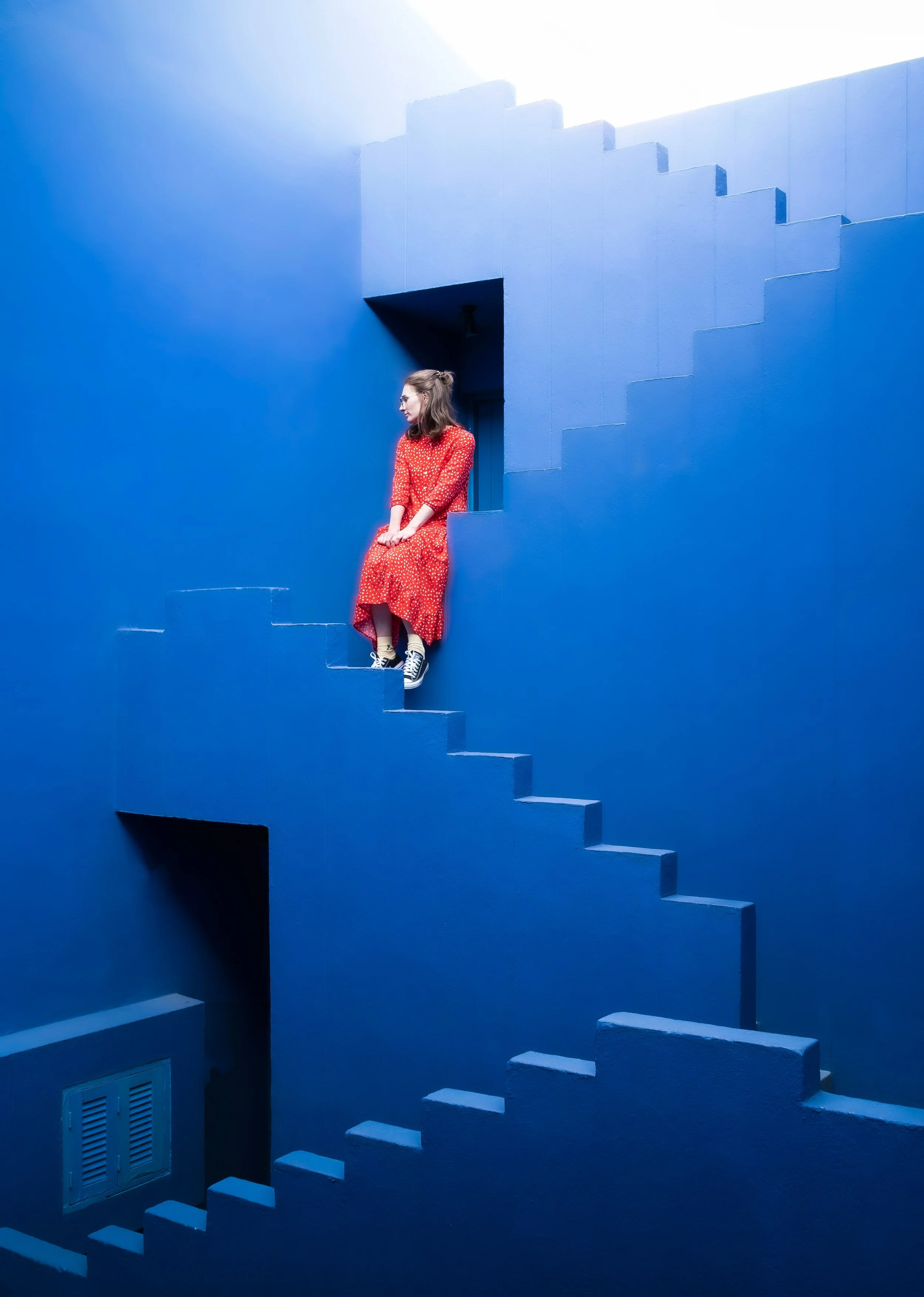 A person in a red polka-dot dress an black sneakers sits amid vivid blue stairways rising in different directions, evoking uncertainty, feeling stuck and searching for a way forward.