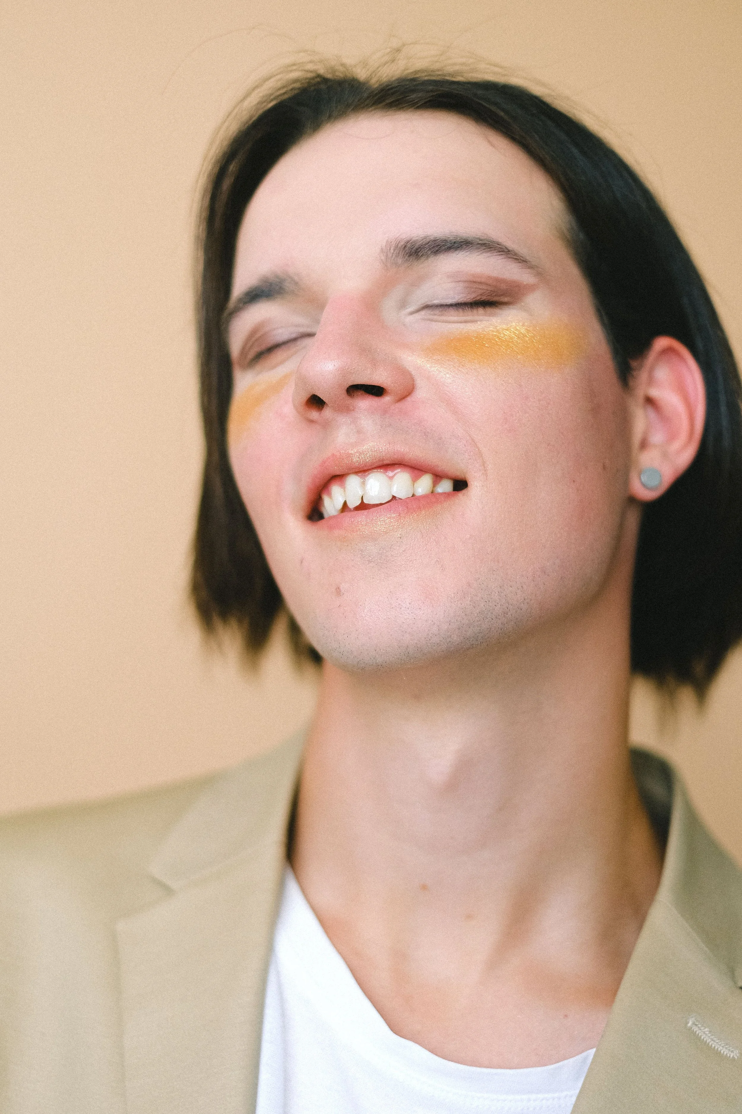 A gender-ambiguous person with short dark hair smiles with their eyes closed, wearing a bright gold stripe of makeup across their cheek.
