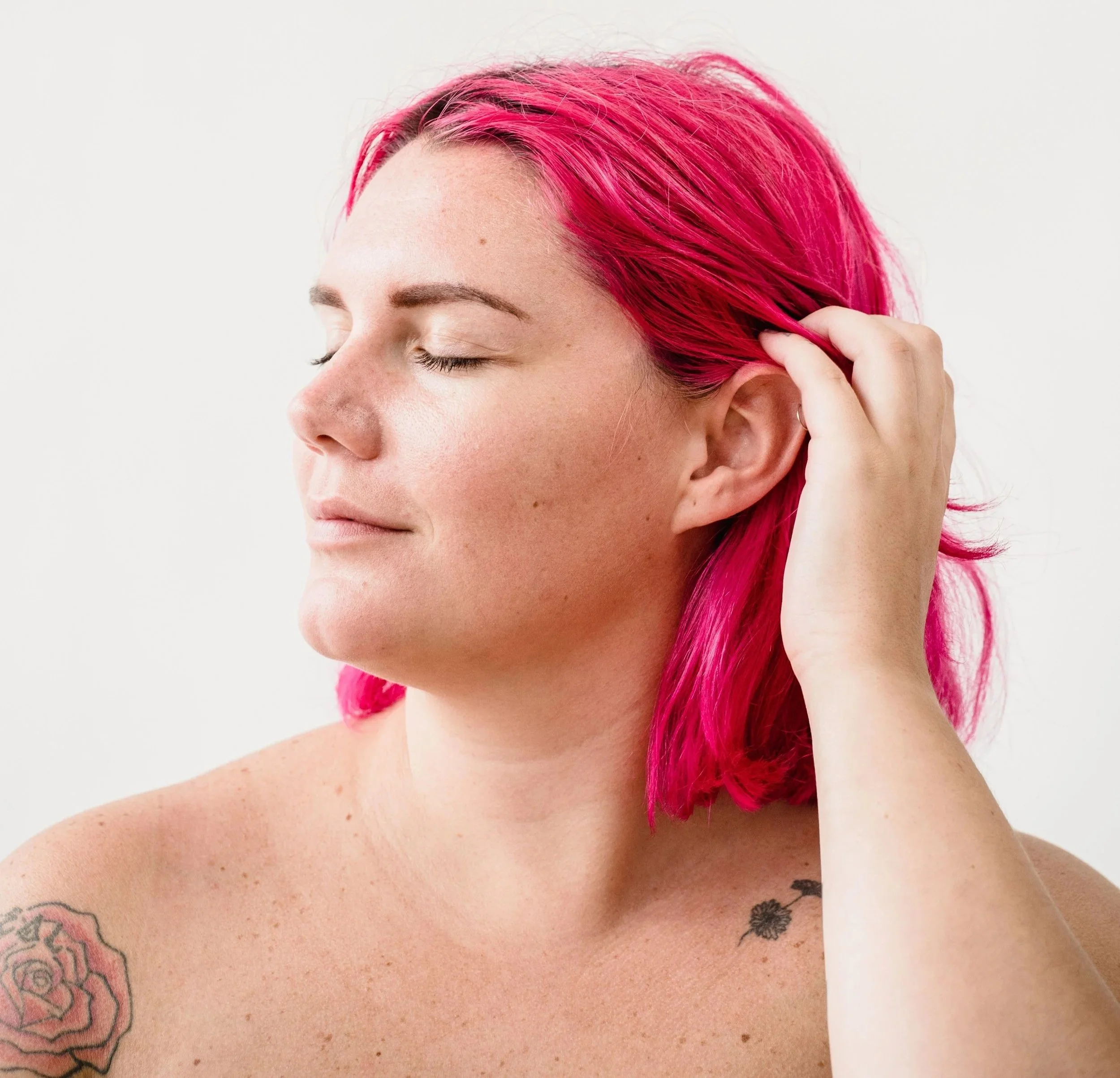 A small-fat white person with vivid pink hair closes their eyes and runs a hand through their hair, appearing calm and absorbed in the pleasure of their own touch.