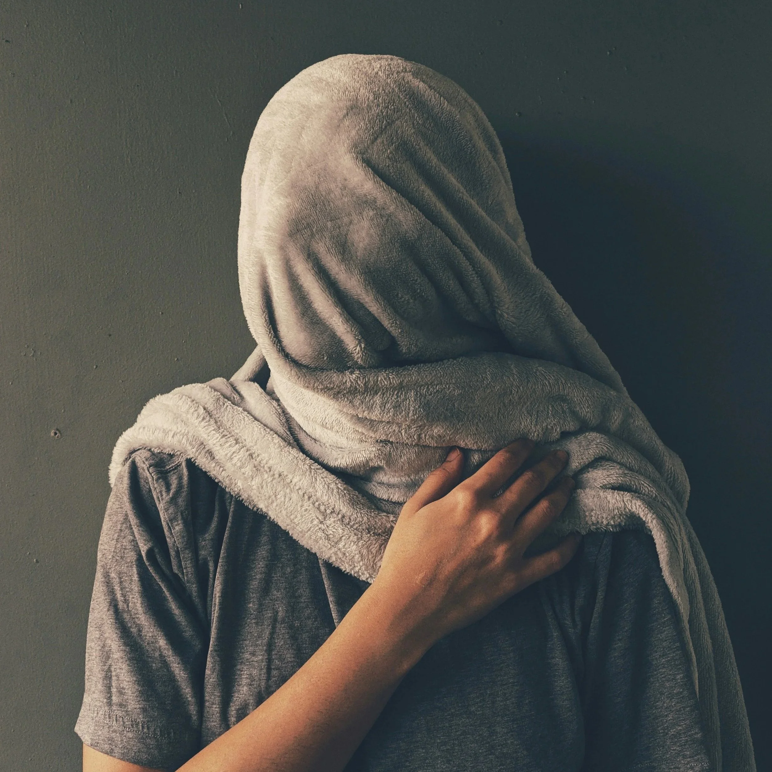 A person of undefined gender has wrapped a grey towel over their head. This evokes feelings of hiding, unhappiness and fatique. They are wearing a grey shirt and are in front of a grey wall.
