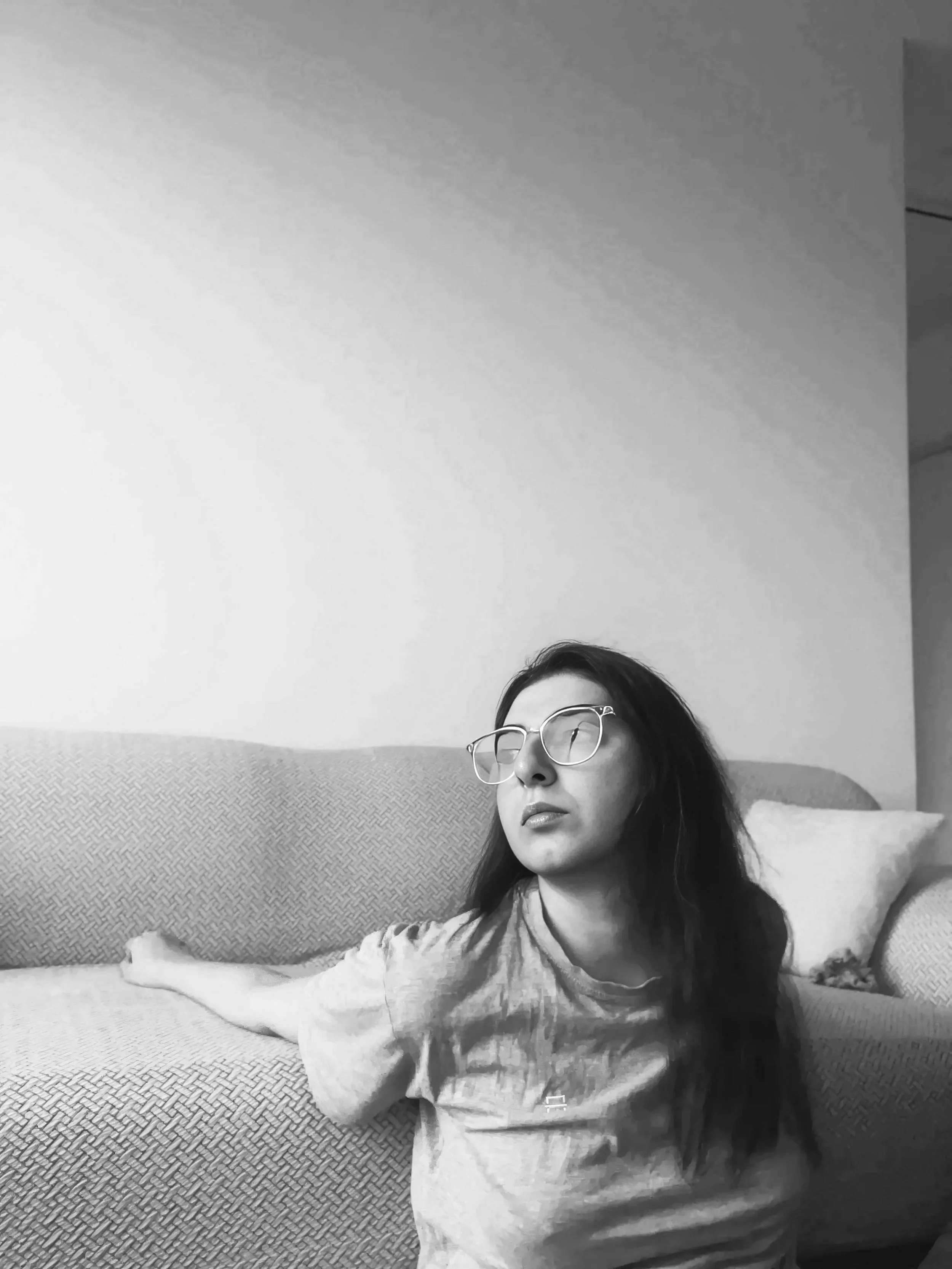 A person wearing glasses reclines on a sofa with their eyes gazing to the side and one arm outstretched.