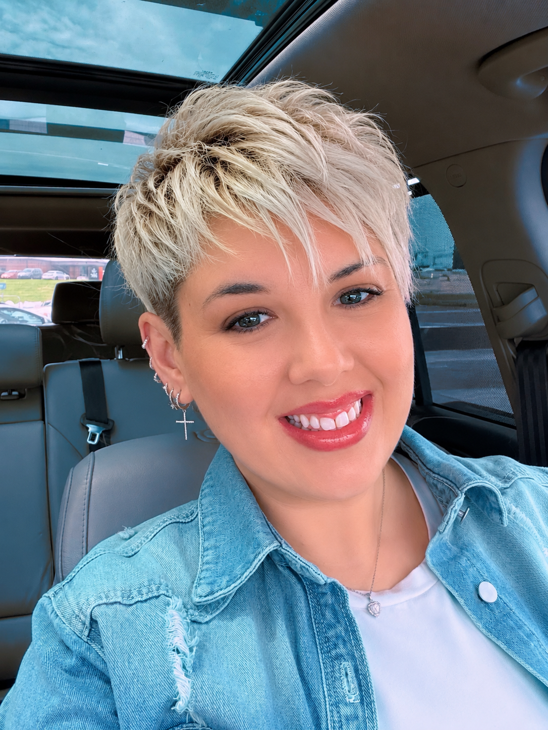 A woman with short, blonde, textured hair smiling inside a car with a sunroof, wearing a denim jacket and earrings.