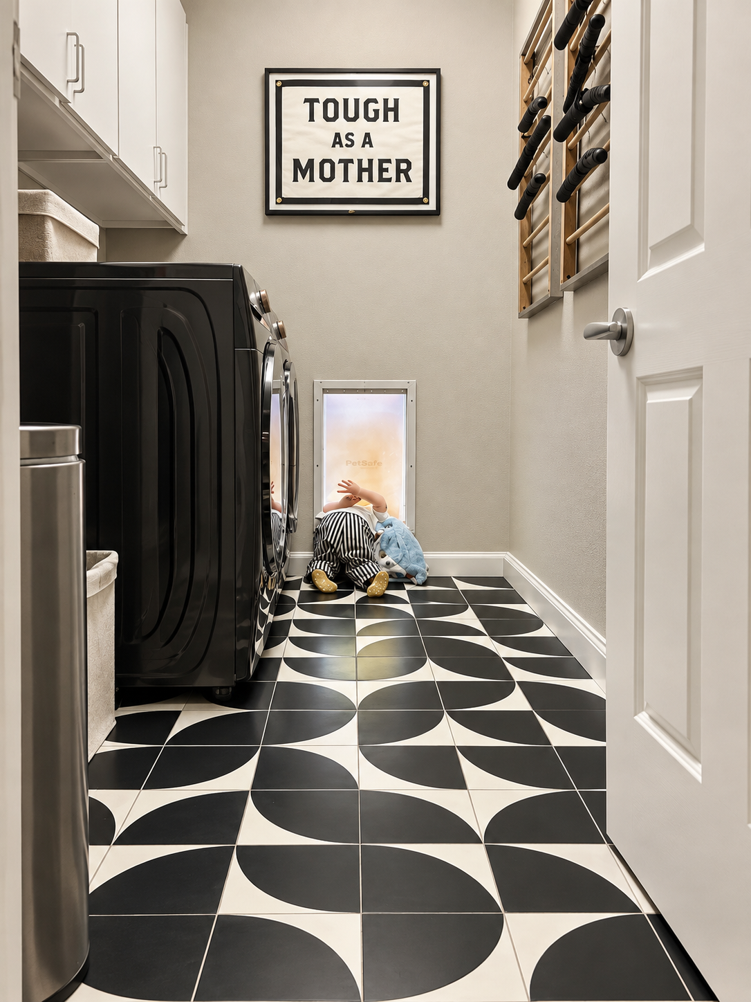 A laundry room with black and white geometric patterned floor tiles, a white cabinet, a framed sign that reads "TOUGH AS A MOTHER," wall-mounted knapsack rack, black washer and dryer, plush toys, and a pet door near the floor.