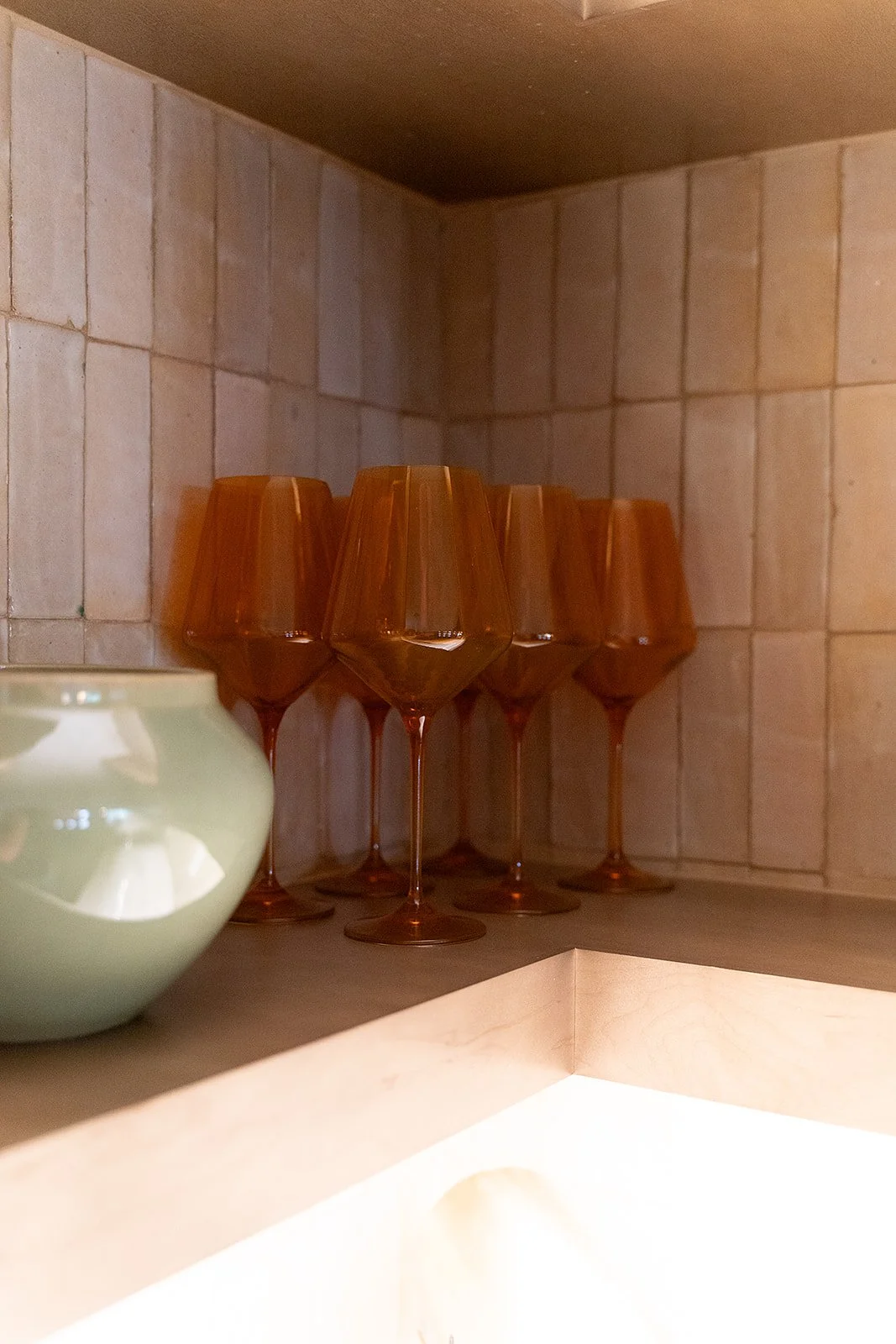 Six amber-colored wine glasses arranged upside down on a kitchen shelf next to a light green ceramic bowl, with a tiled wall in the background.