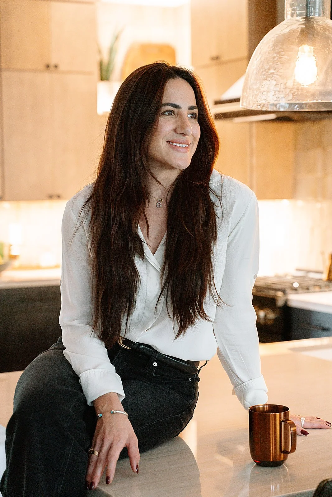 A woman with long dark hair, smiling, in a kitchen with warm lighting, wearing a white blouse and black pants, sitting on a kitchen counter next to a copper-colored mug.
