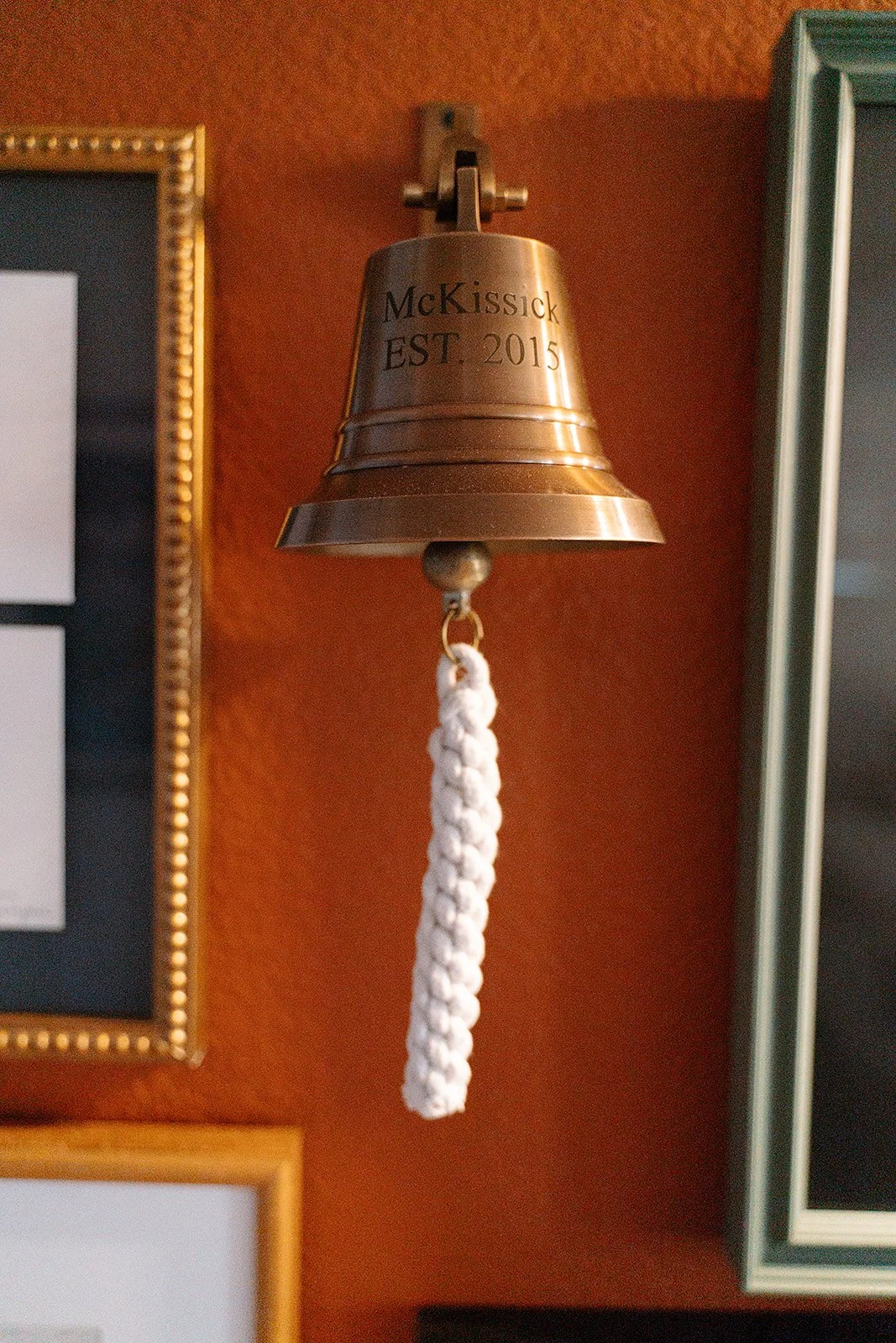 A metal bell with the inscription "McKissick EST. 2015" hangs on a wall with a white rope attached to its clapper.
