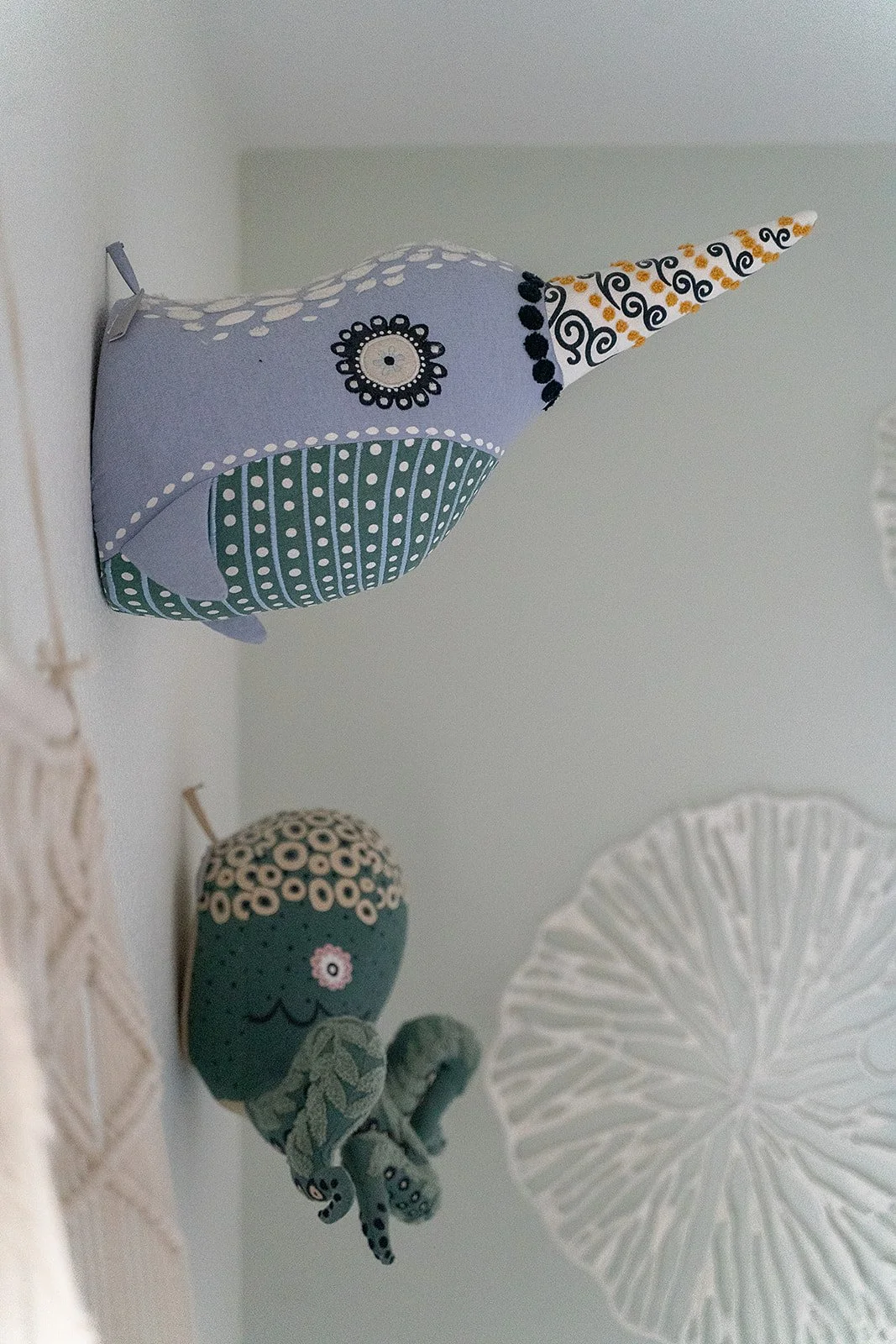 Decorative stuffed sea creature resembling a narwhal with a long, spiral horn, polka dots, and lace patterns, mounted on a wall with roof and ceiling in background.