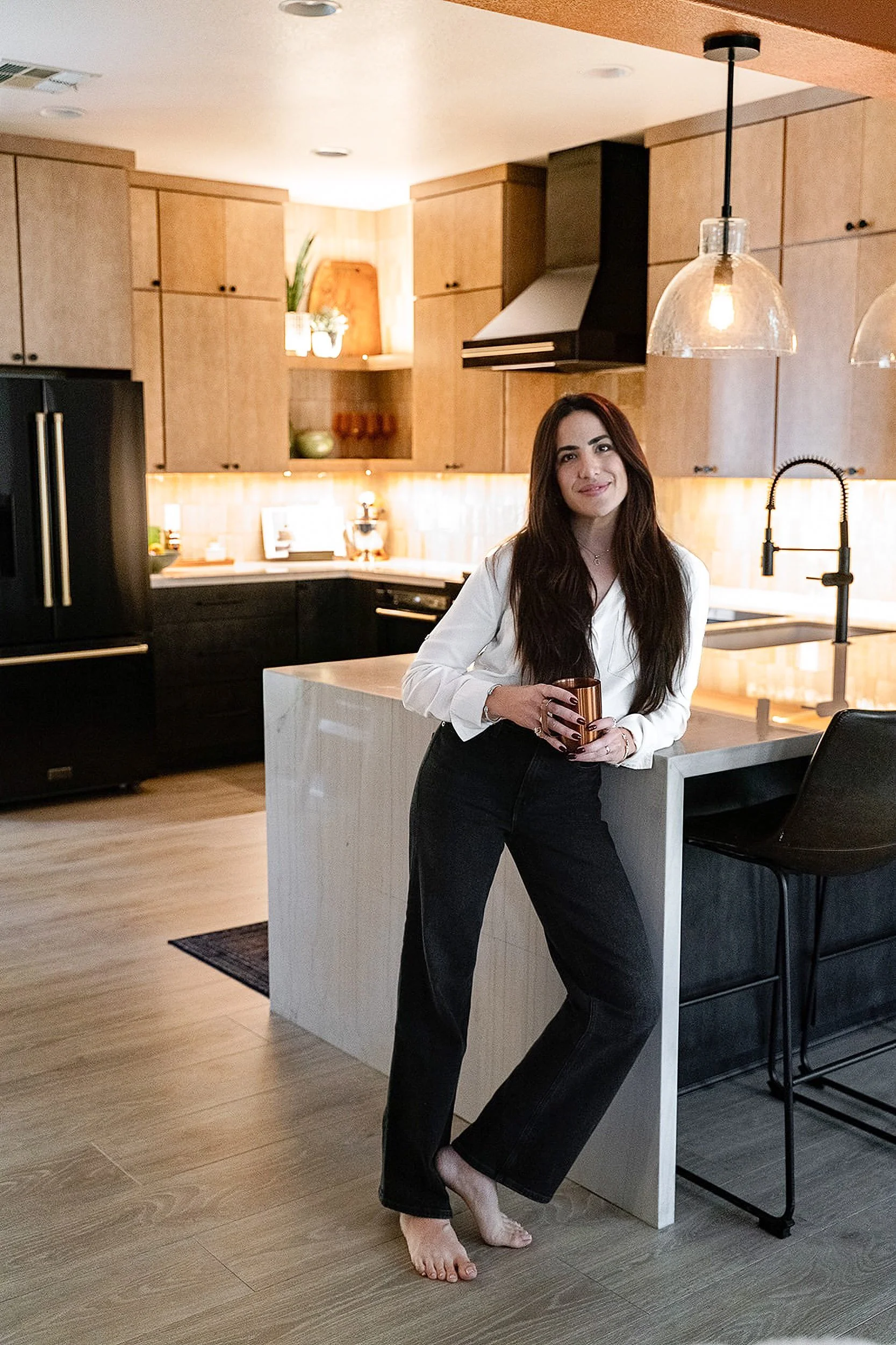A woman with long dark hair, wearing a white blouse and black pants, stands barefoot in a modern kitchen. She is holding a copper mug and smiling at the camera. The kitchen has wooden cabinets, a black refrigerator, and a marble island with a black chair. There are pendant lights hanging from the ceiling.