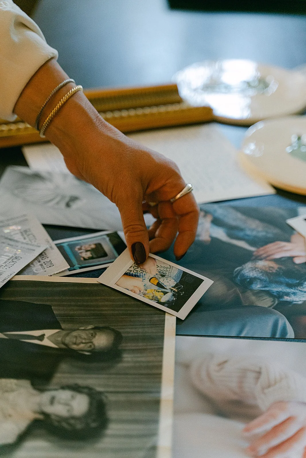 A person's hand with bracelets and rings picking up a small photograph from a table covered with various photos and paper memorabilia.