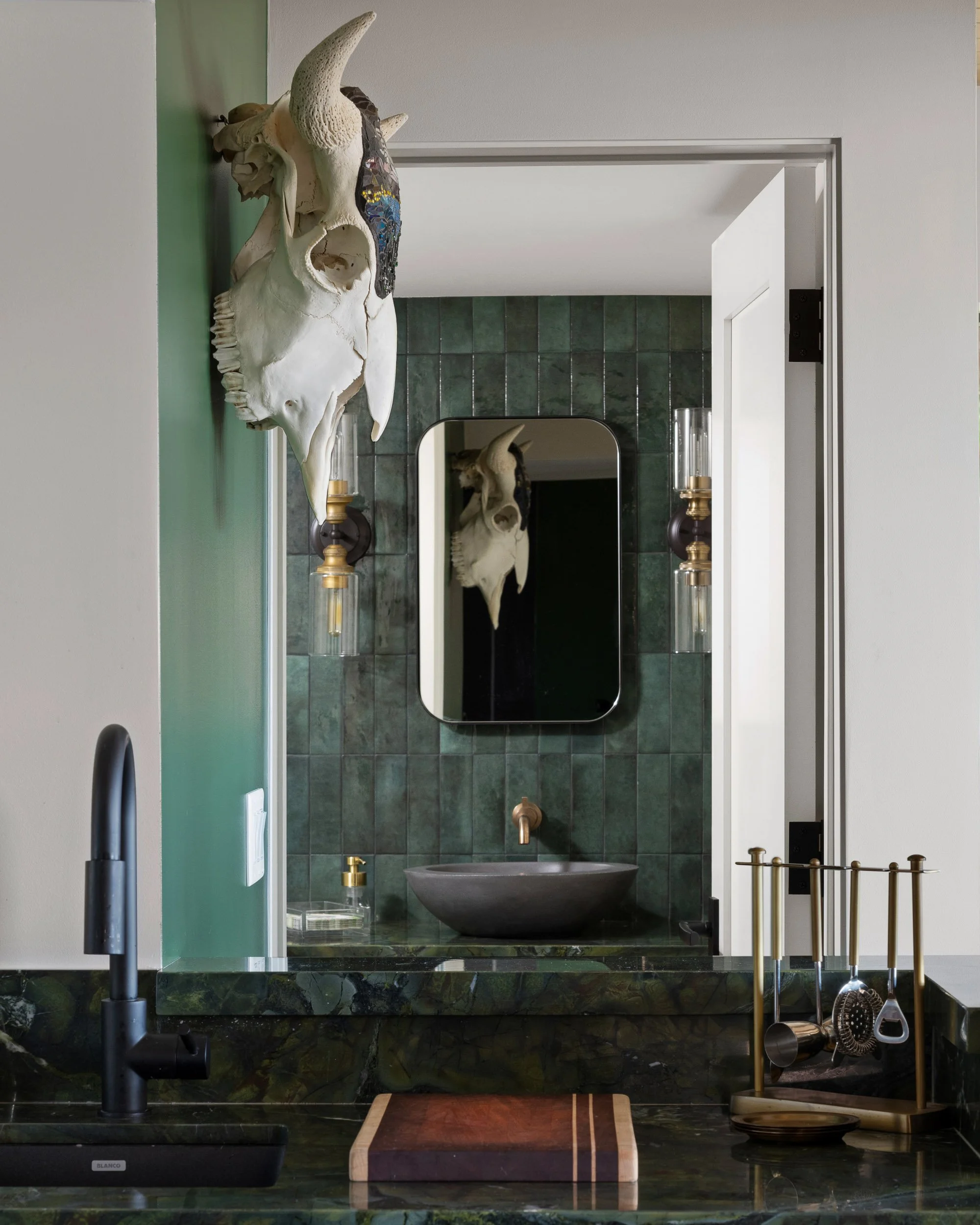 A kitchen or bar area with a green tiled wall, a large skull mounted on the wall, and a mirror reflecting the skull. There is a black sink with a modern black faucet, a cutting board with a dark wood top and a light wood border, and a brass and glass light fixture on the wall. On the right side, a brass and black utensil holder with cooking utensils is visible.