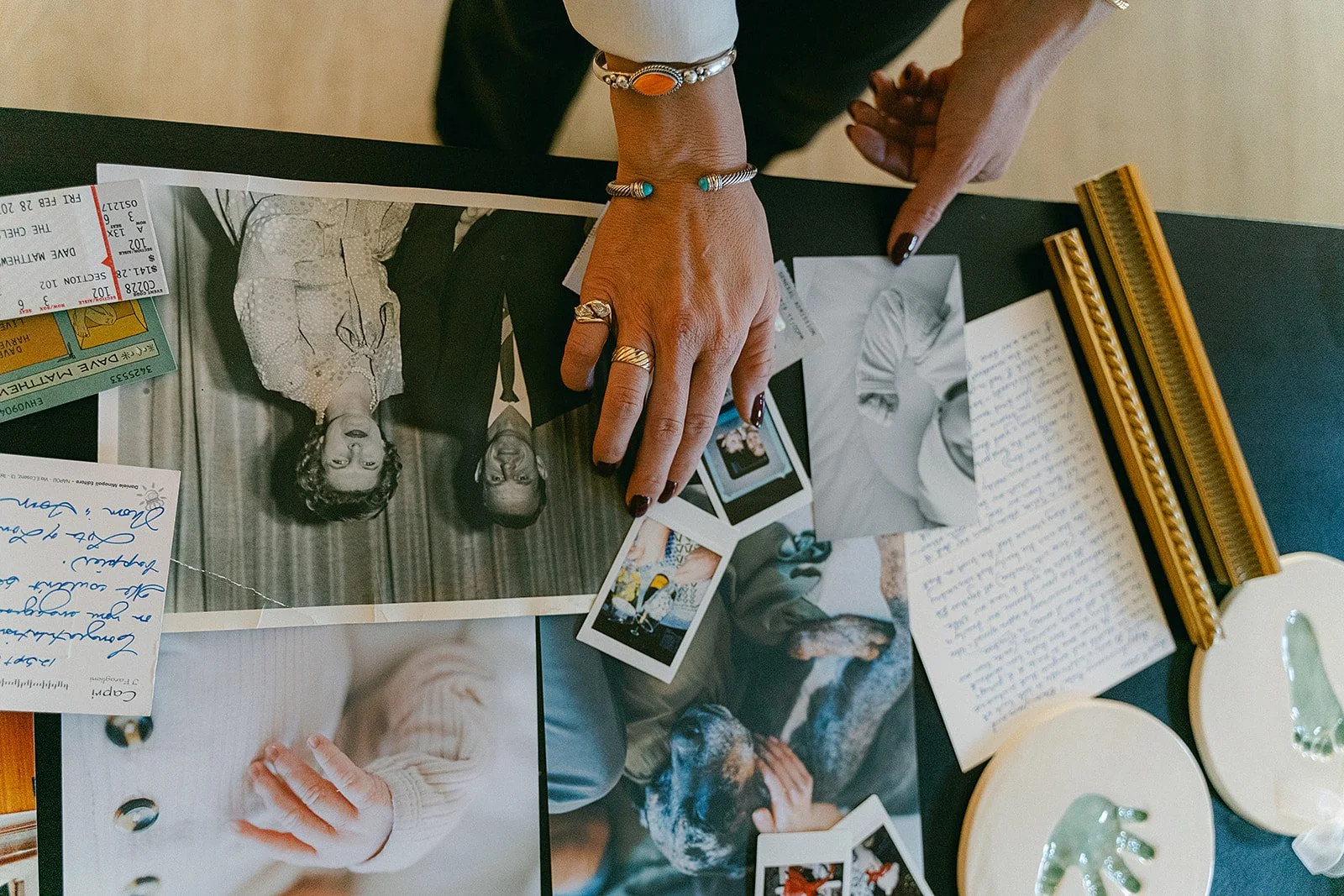 Close-up of two hands with jewelry pointing to black-and-white photographs, surrounded by handwritten letters, printed images, and a framed photo, on a black table.
