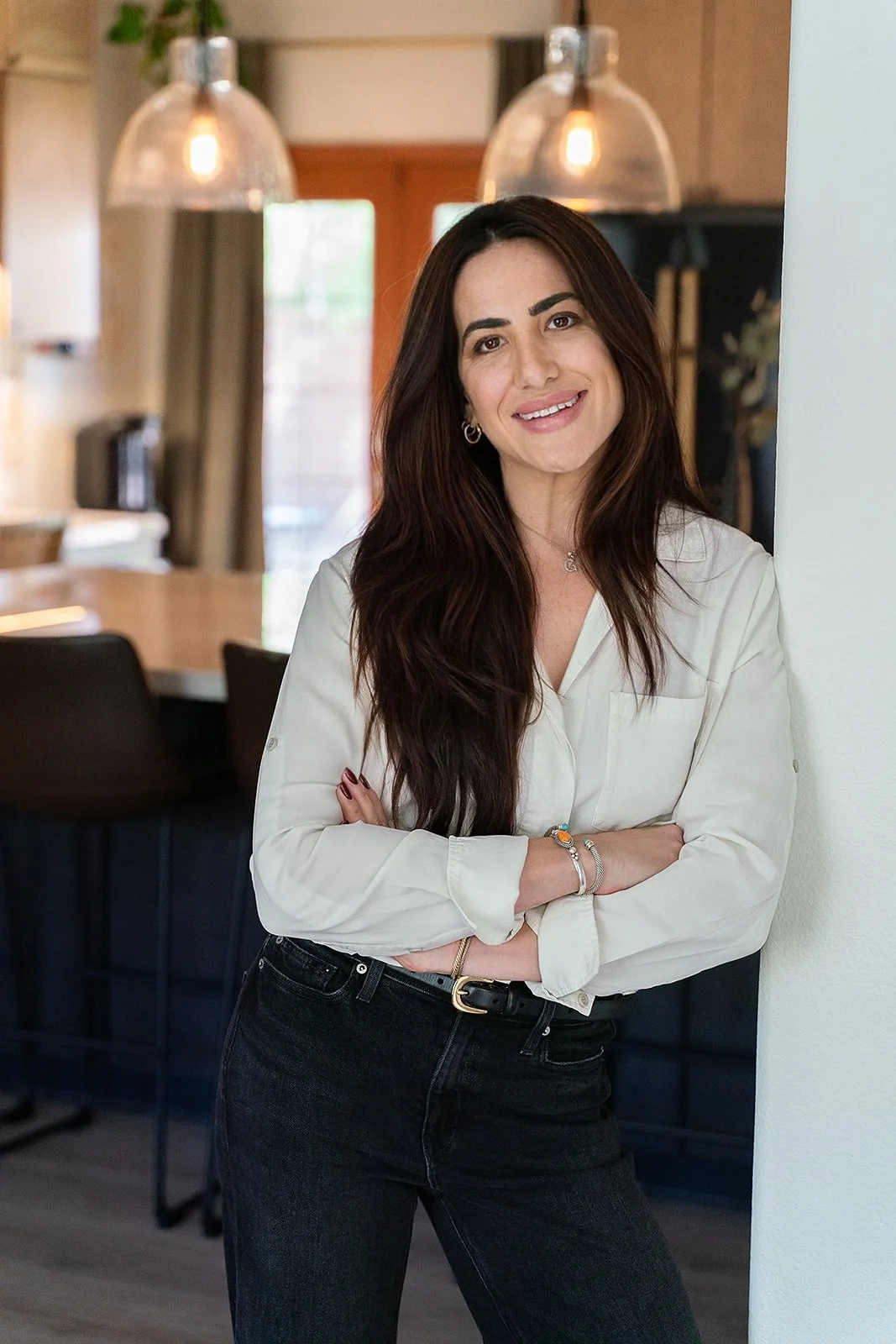 A woman with long dark hair, wearing a white blouse and black jeans, standing in a modern kitchen with pendant lights and a dining area in the background.