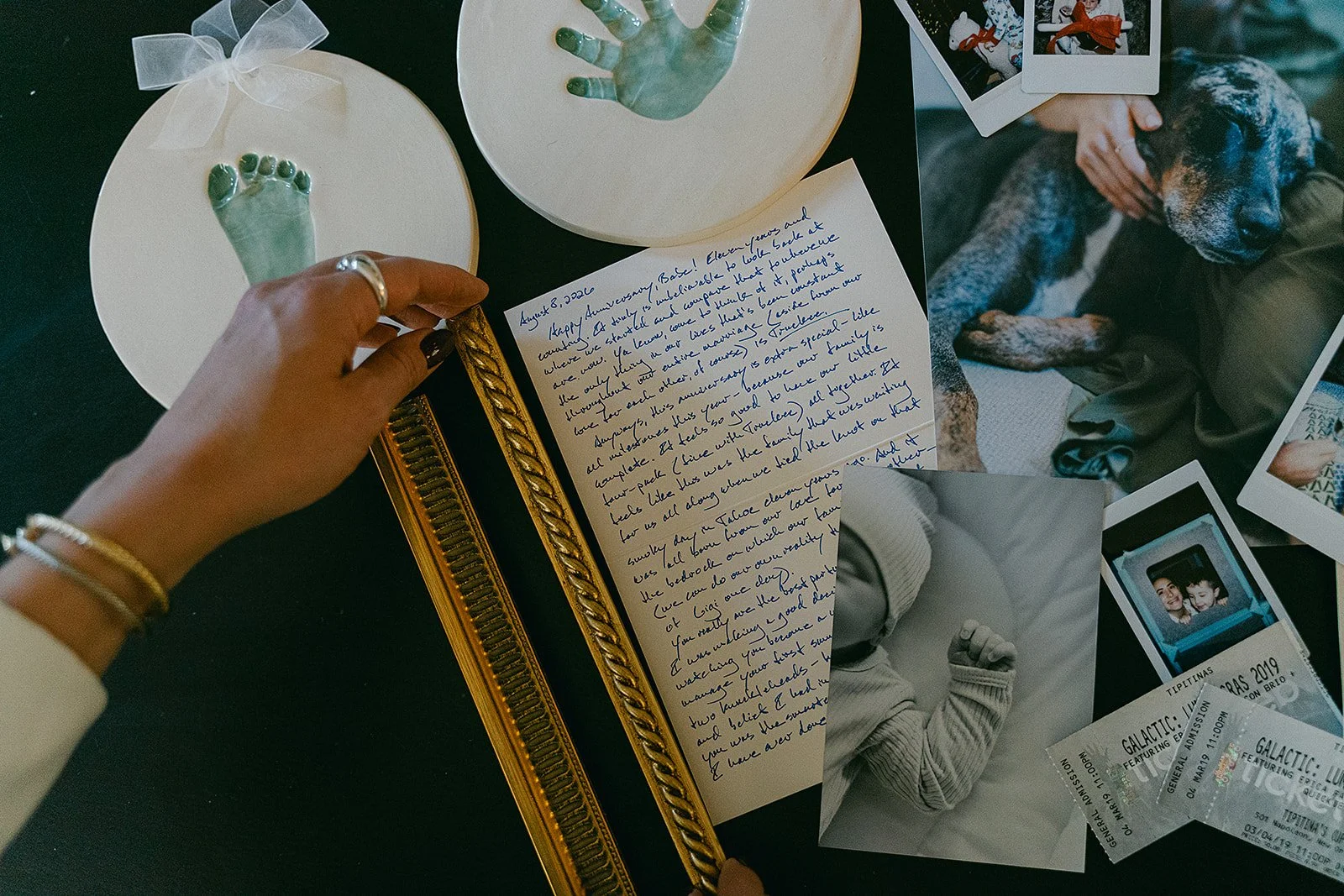 A table with framed handprints, a handwritten letter, Polaroid photos of a dog and a person, event tickets, and various personal items.