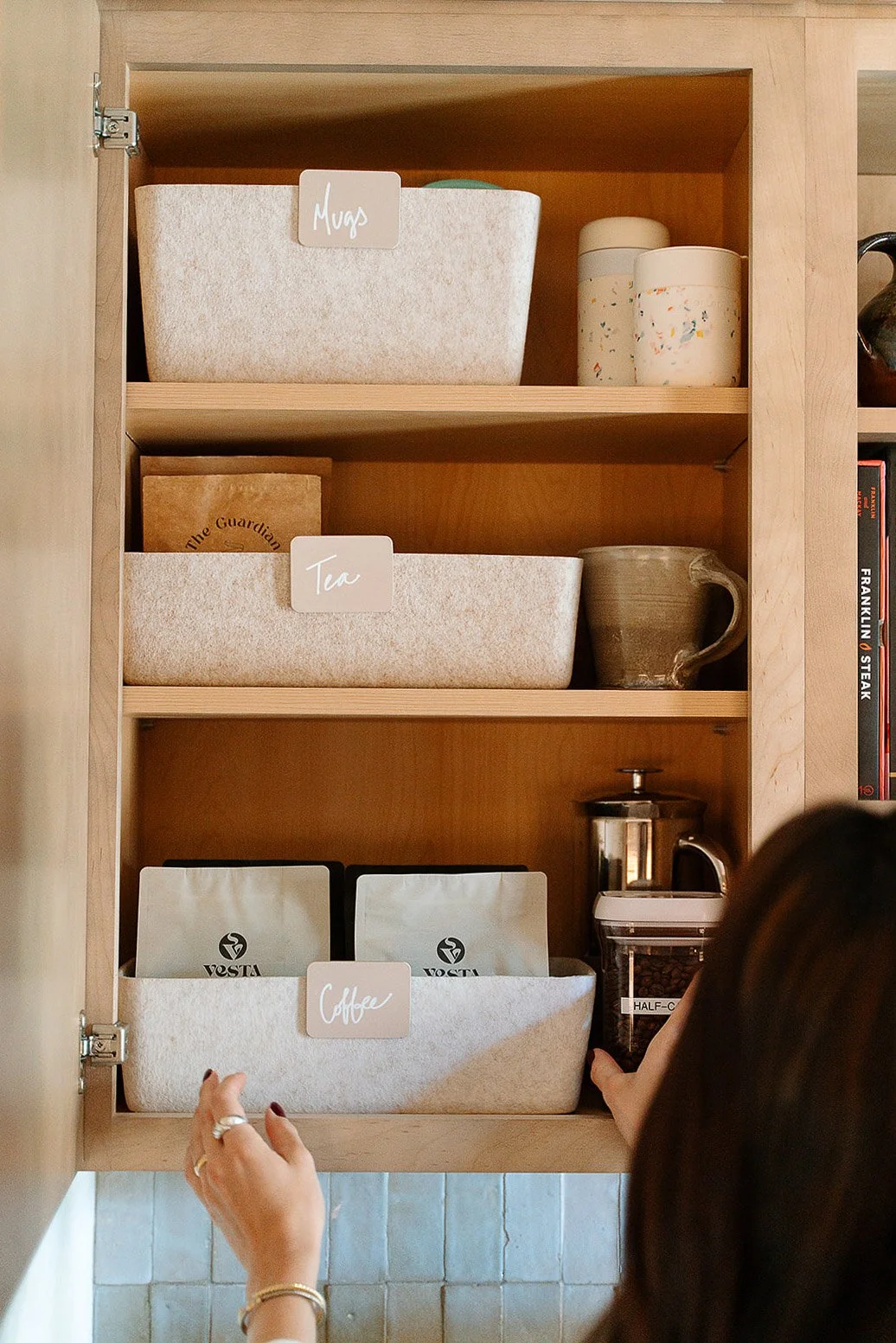 Open wooden kitchen cabinet with labeled storage bins for mugs, tea, and coffee. Someone is reaching for coffee beans from a container.