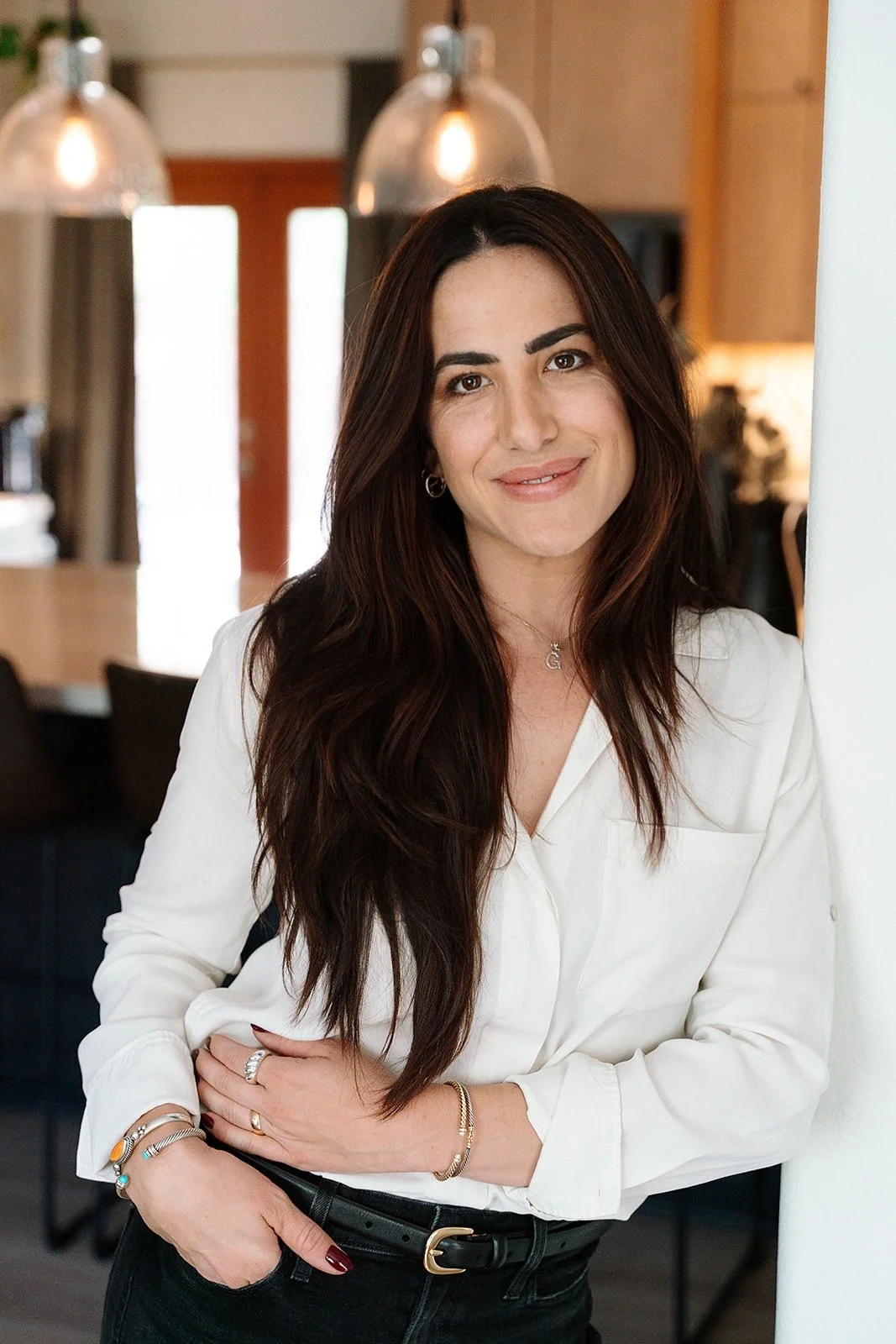 A woman with long, dark brown hair, smiling, wearing a white blouse, multiple jewelry bracelets, rings, and a necklace, standing in a cozy, modern indoor space with warm lighting and wooden accents.