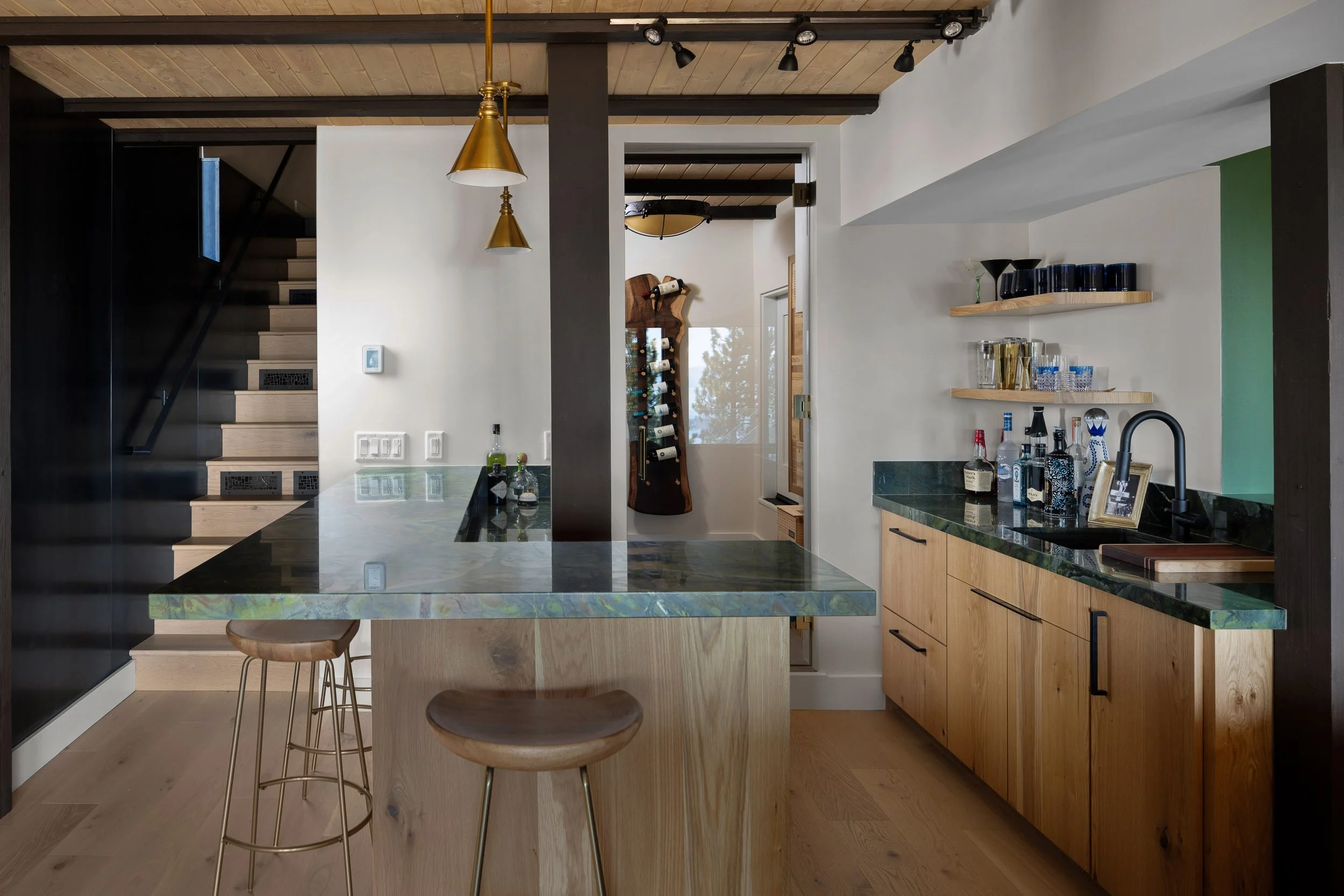 Modern kitchen with wooden cabinets, a green marble countertop, and open shelves holding glasses and bottles. A black faucet is visible at the sink, and there are bar stools at the island. The ceiling has wood paneling with track lighting, and a doorway leads to a room with a wine rack.